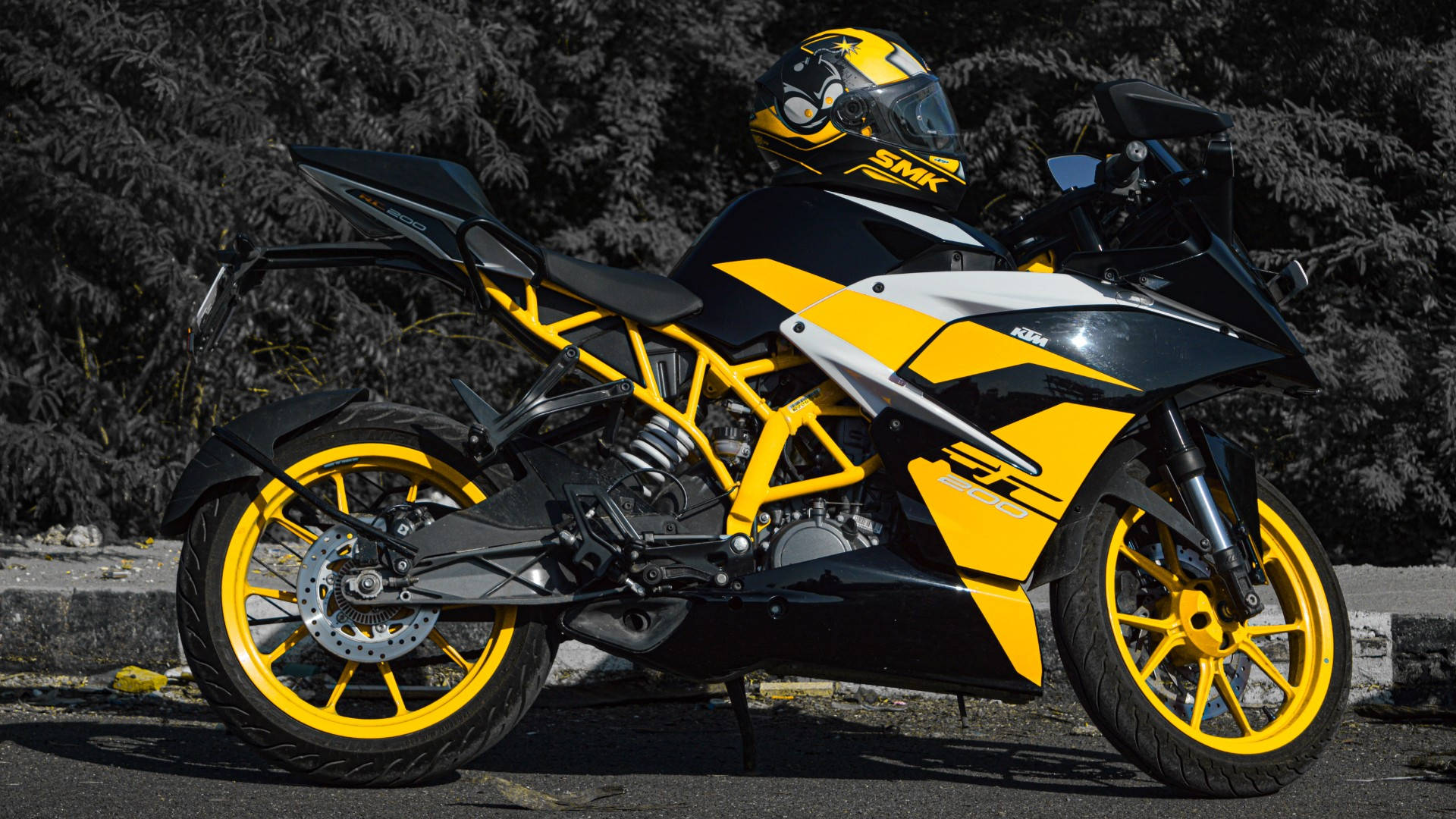 酷炫黃色KTM RC 200 壁紙 — 一架搶眼的黃黑配色運動電單車，連同一個相配的頭盔，停泊在鋪砌道路上，旁邊有深色常綠樹。