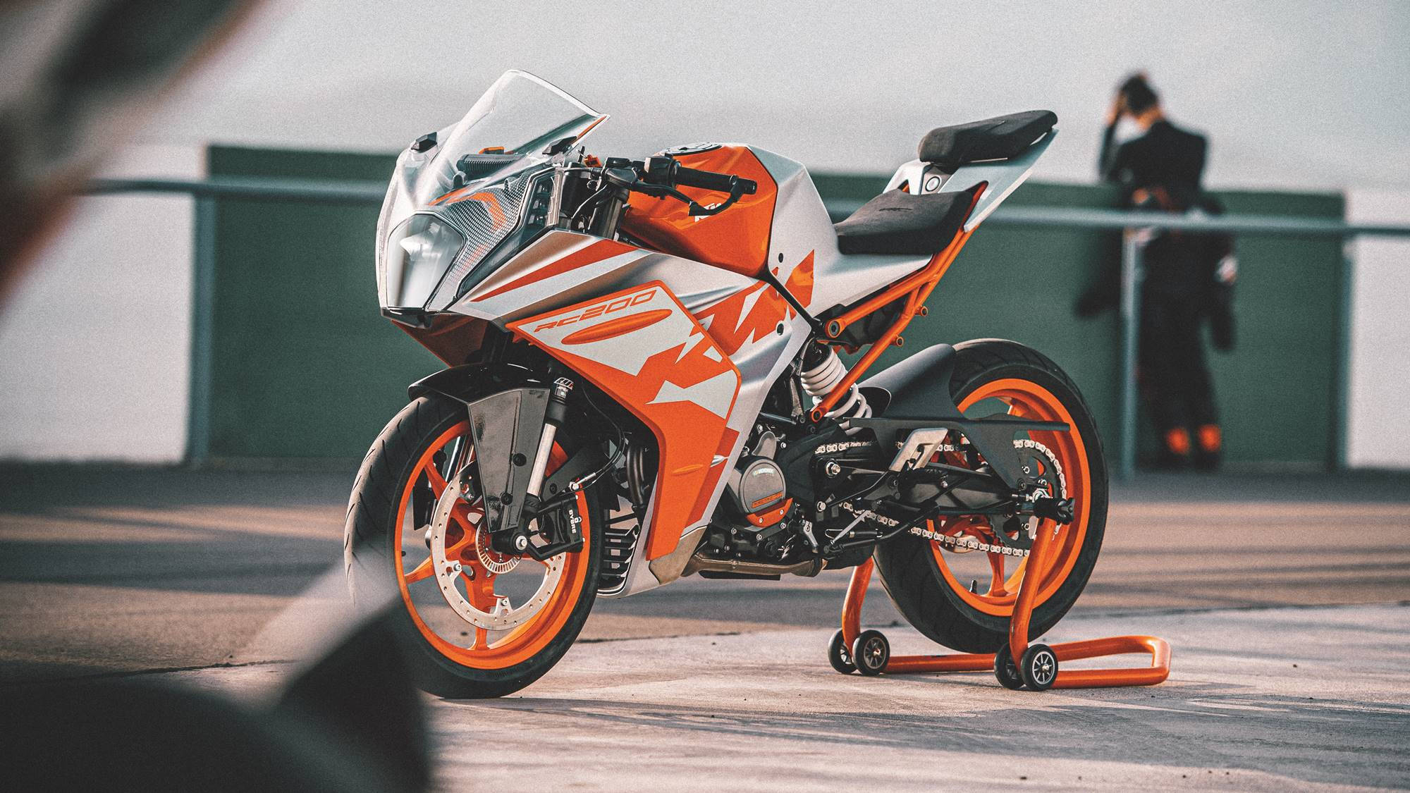 賽道上的KTM RC 200 壁紙 — 一架時尚的橙色和白色KTM RC200運動電單車放在混凝土平台上的支架上，背景有一個模糊的人影。