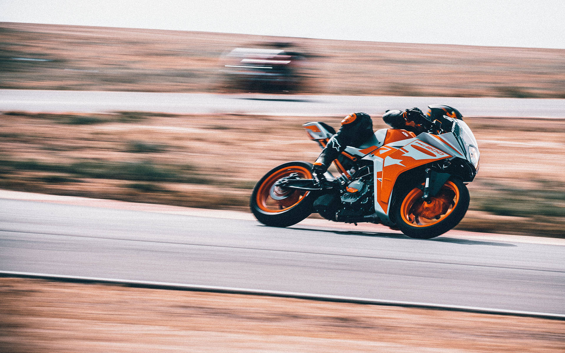 KTM RC 200轉彎 壁紙 — 一名騎手騎著橙白色運動電單車，在沙漠公路上高速飛馳，並帶有動態模糊效果地傾斜入彎。