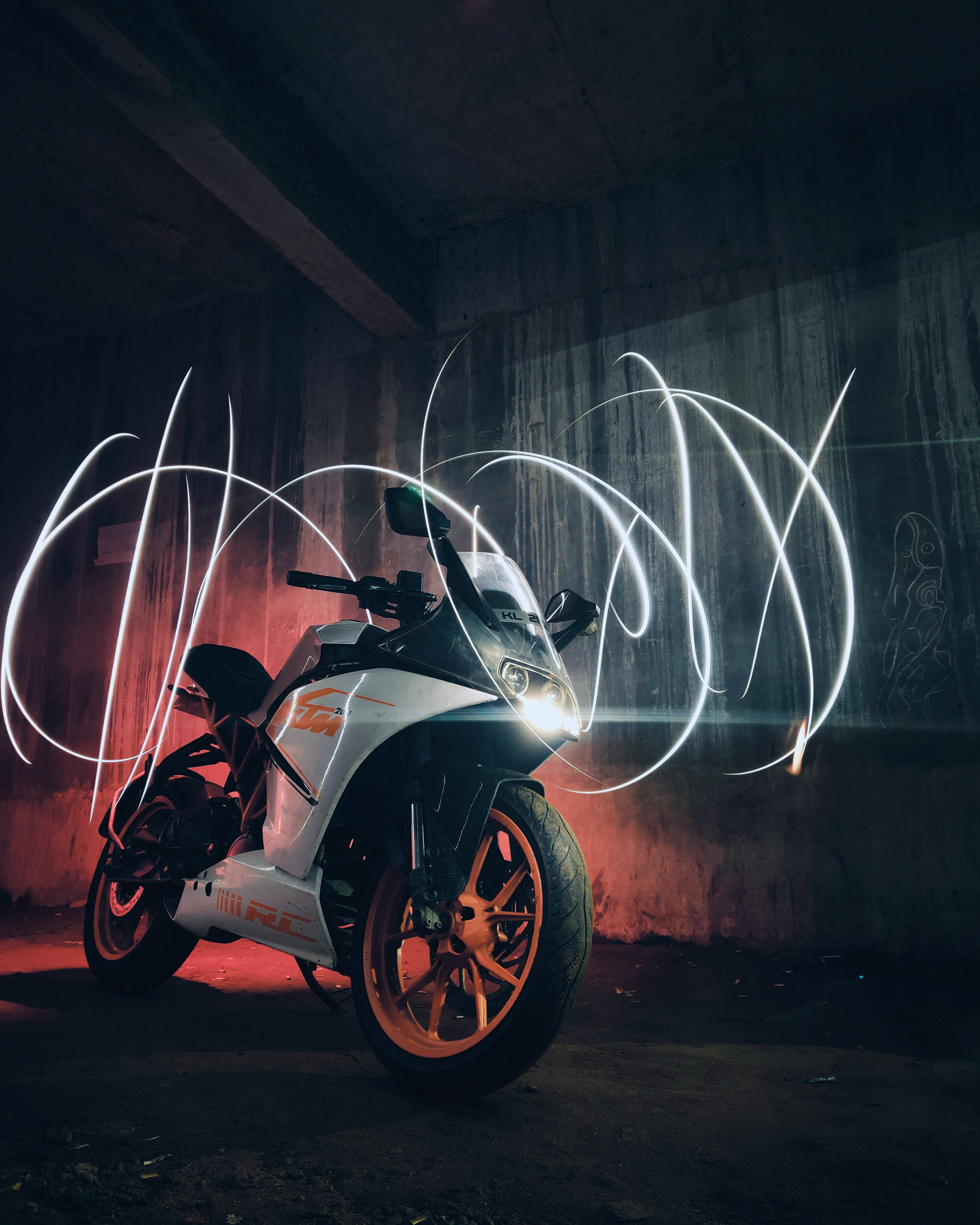 KTM RC 390 2016 光影遊戲 壁紙 — 一輛白色和橙色的KTM RC電單車在粗糙的混凝土隧道中，在戲劇性的紅色燈光和旋轉的白色光軌下閃閃發光。