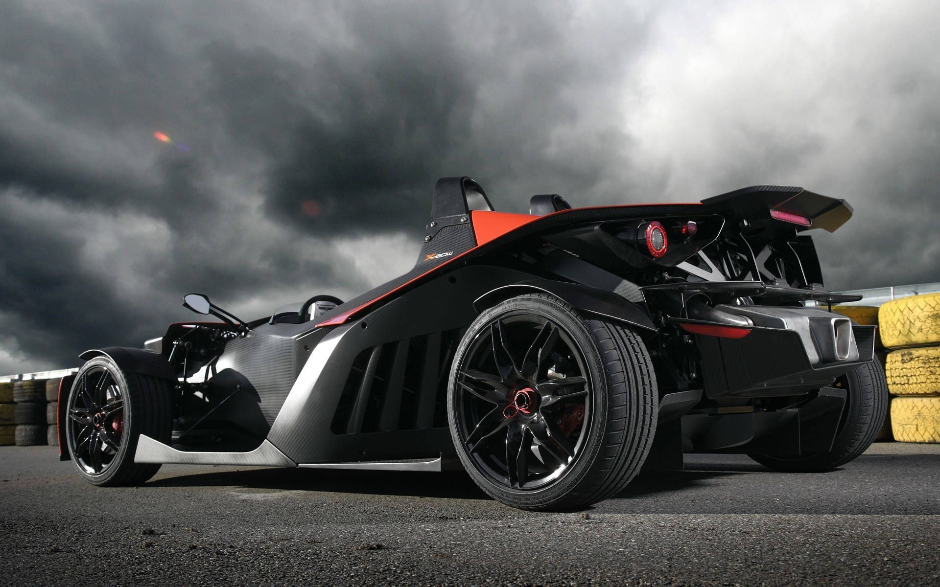 KTM X-Bow 最佳桌面壁紙 壁紙 — 一輛時尚的黑色和橙色跑車，帶有碳纖維細節，在暴風雨的天空下停在賽道上。