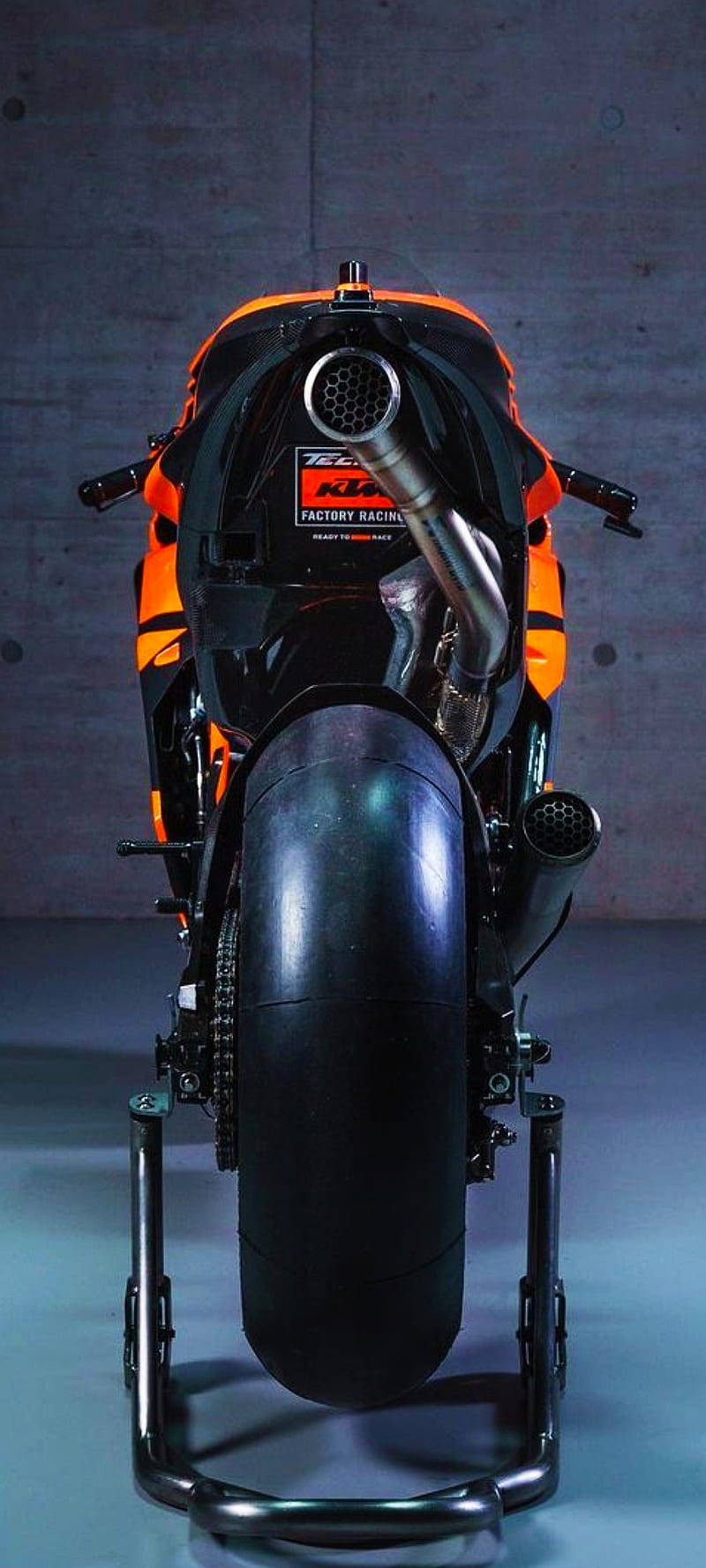 KTM摩托車iPhone背面視圖 壁紙 — 一輛時尚的橙色和黑色KTM Factory Racing電單車放在支架上，背景是混凝土牆的後視圖。