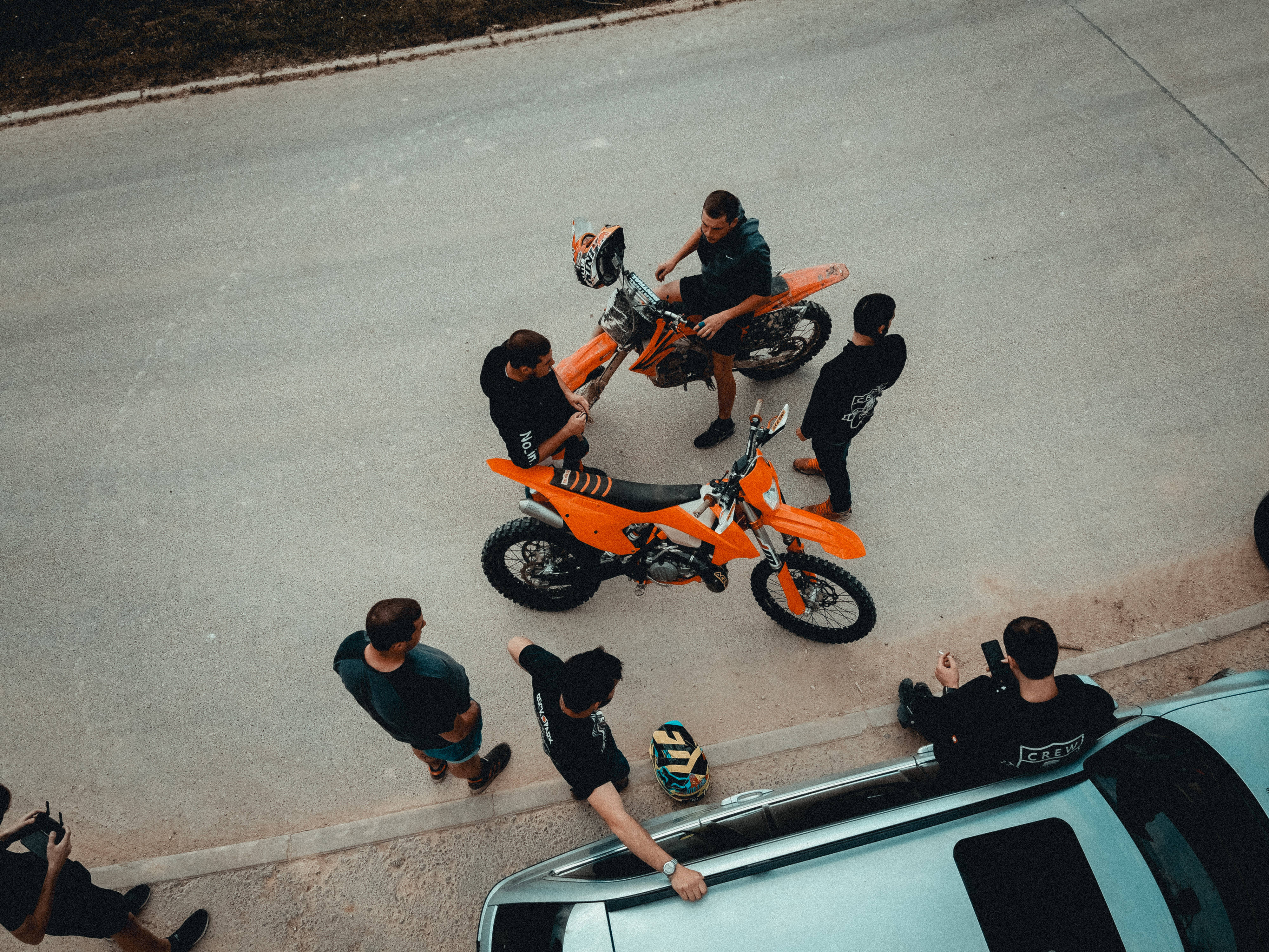KTM機車手休息 壁紙 — Overhead view of a group of men gathered around two bright orange dirt bikes on a paved road beside a silver car.