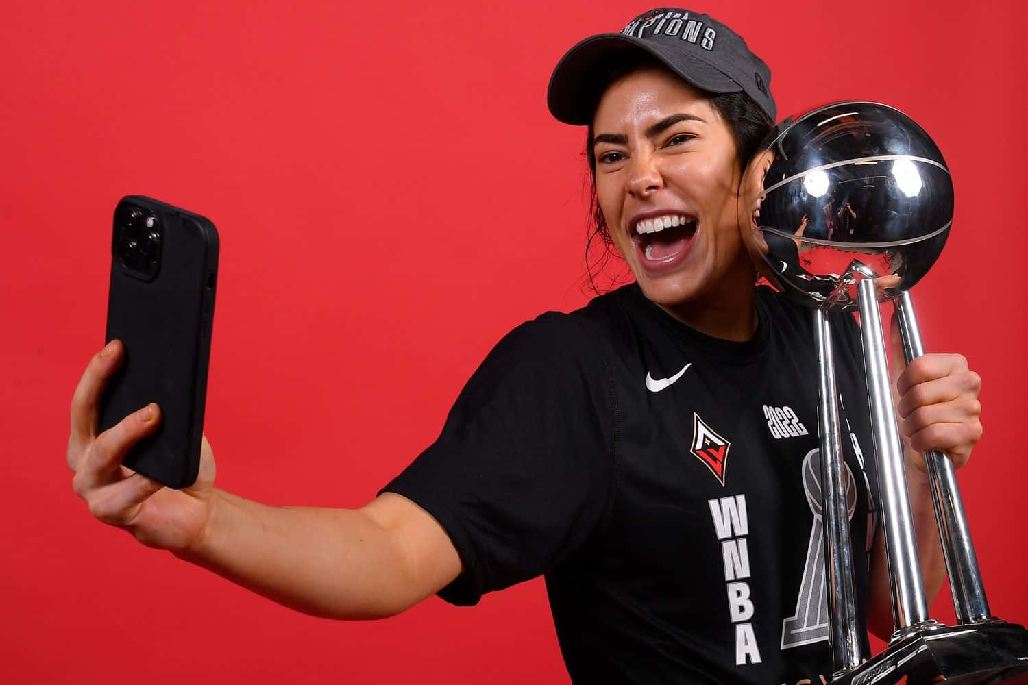  壁紙 — 一位快樂的女籃球員，戴著黑色 'Champions' 帽子和球衣，手持閃亮的 WNBA 獎盃，在鮮紅色的背景下自拍，笑容燦爛。
