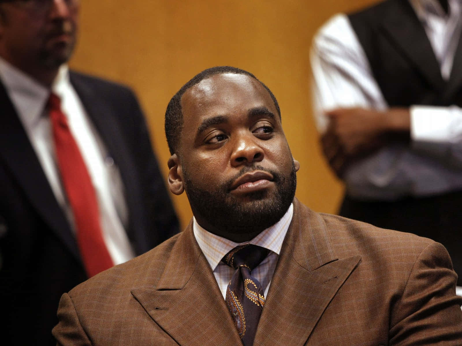 Kwame Kilpatrick出席記者會 壁紙 — 一名身穿棕色西裝及圖案領帶的男子，身後有模糊的人物，莊嚴地坐在法庭內。