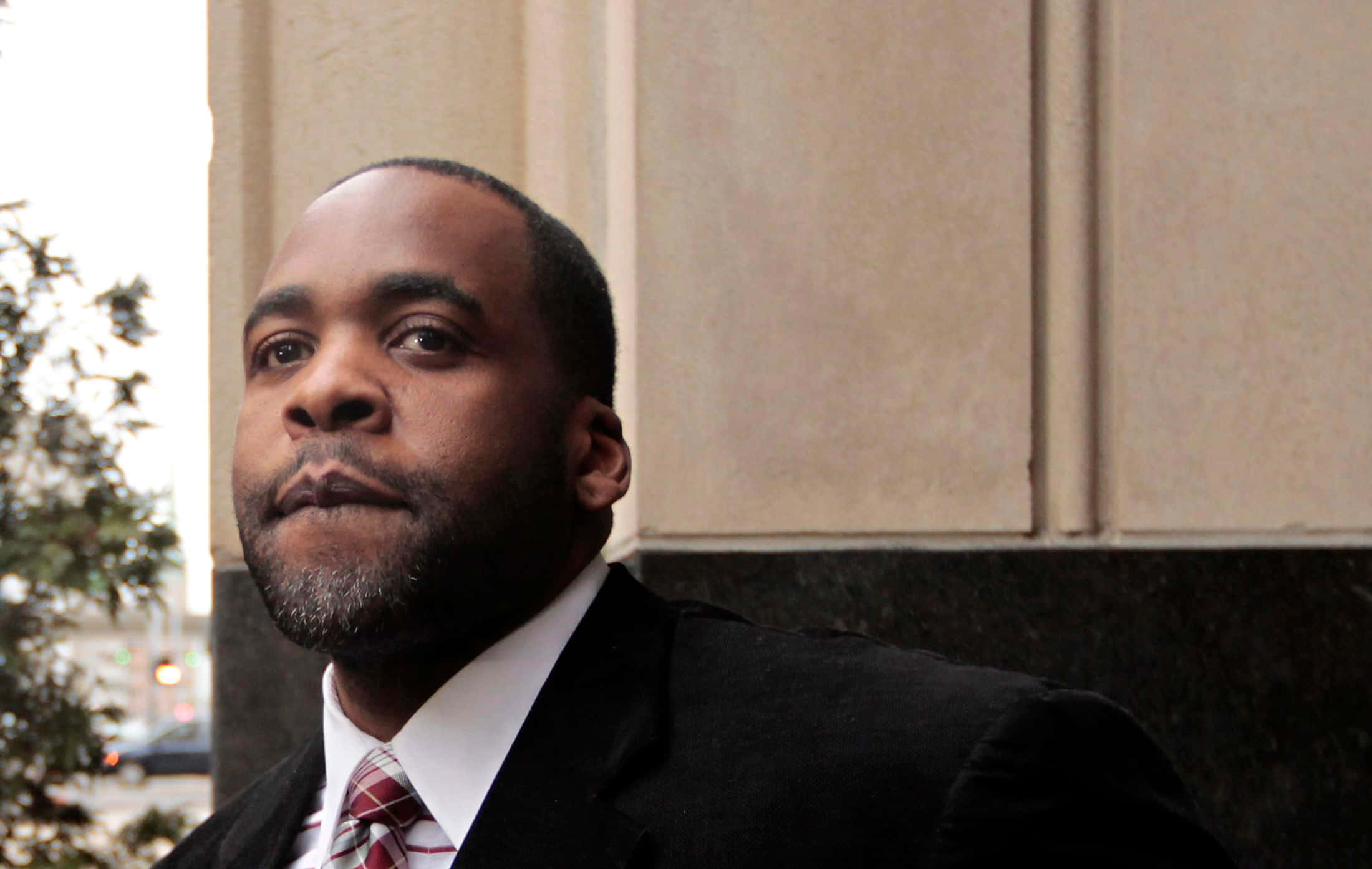 Kwame Kilpatrick身著正裝的官方肖像照 壁紙 — 一個穿著黑色西裝和紅色領帶的嚴肅男子，站在石頭建築前，背景有綠色樹葉。