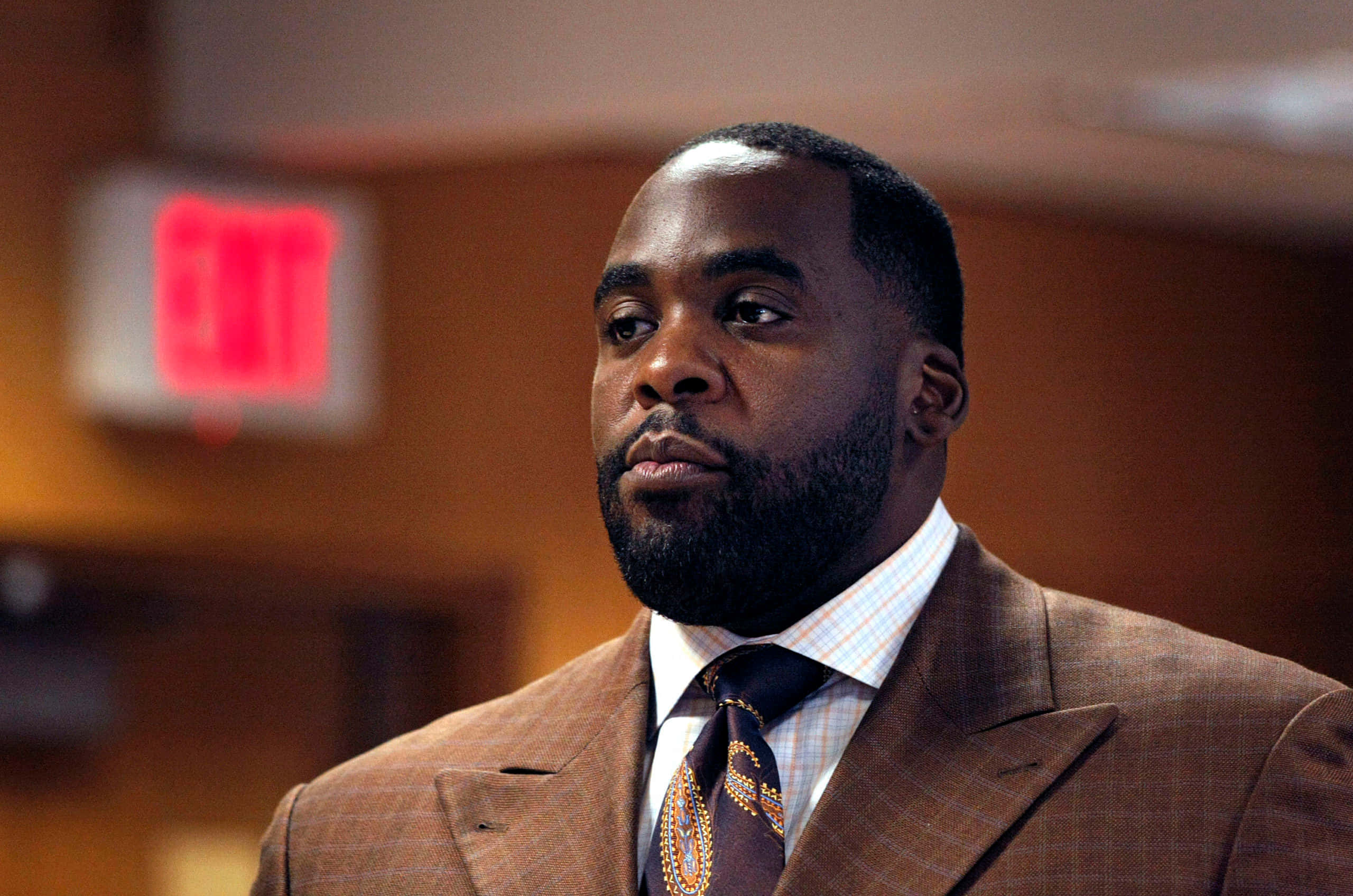 Kwame Kilpatrick出席公開活動 壁紙 — 一個穿著棕色西裝和圖案領帶的嚴肅男子，在溫暖照明的室內環境中看向鏡頭外，背景中有一個模糊的紅色出口標誌。