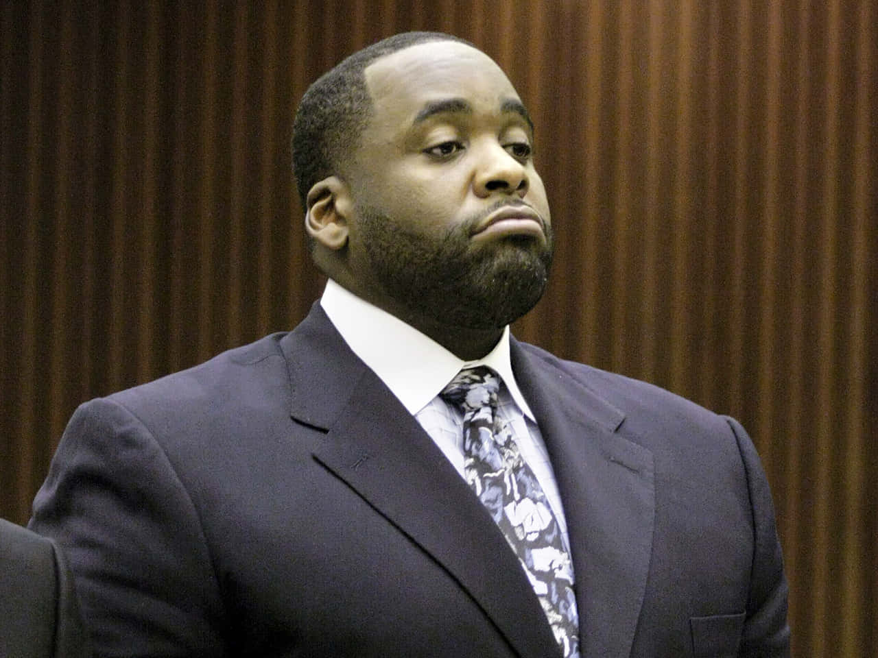 Kwame Kilpatrick公開演講 壁紙 — 一名身穿深色西裝及圖案領帶的男子，在木牆板的法庭內神情嚴肅地向上望。