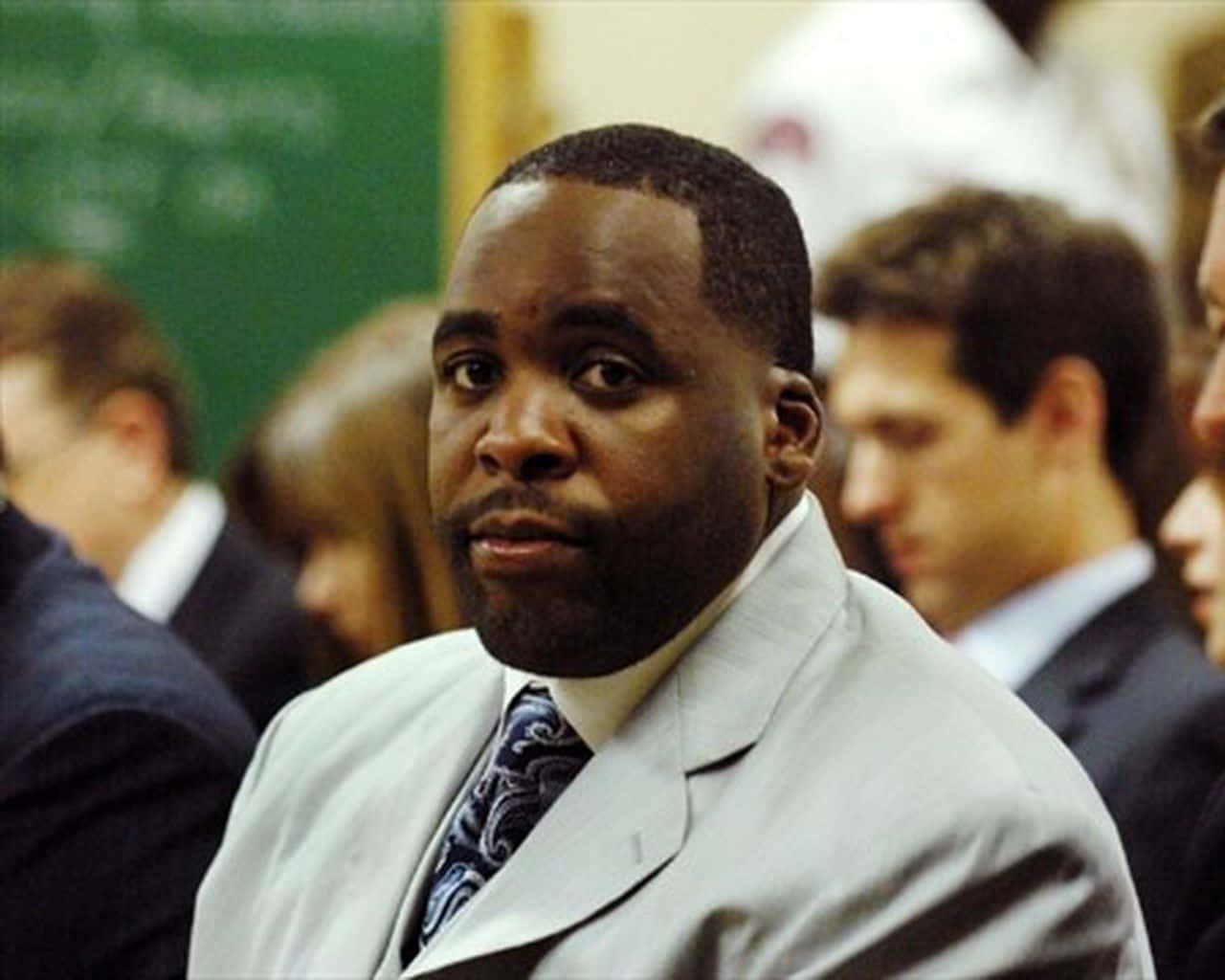 Kwame Kilpatrick出席公開活動 壁紙 — 一名身穿淺灰色西裝和圖案領帶的男子在擁擠的室內環境中，身後有模糊的人群，專注地向前看。