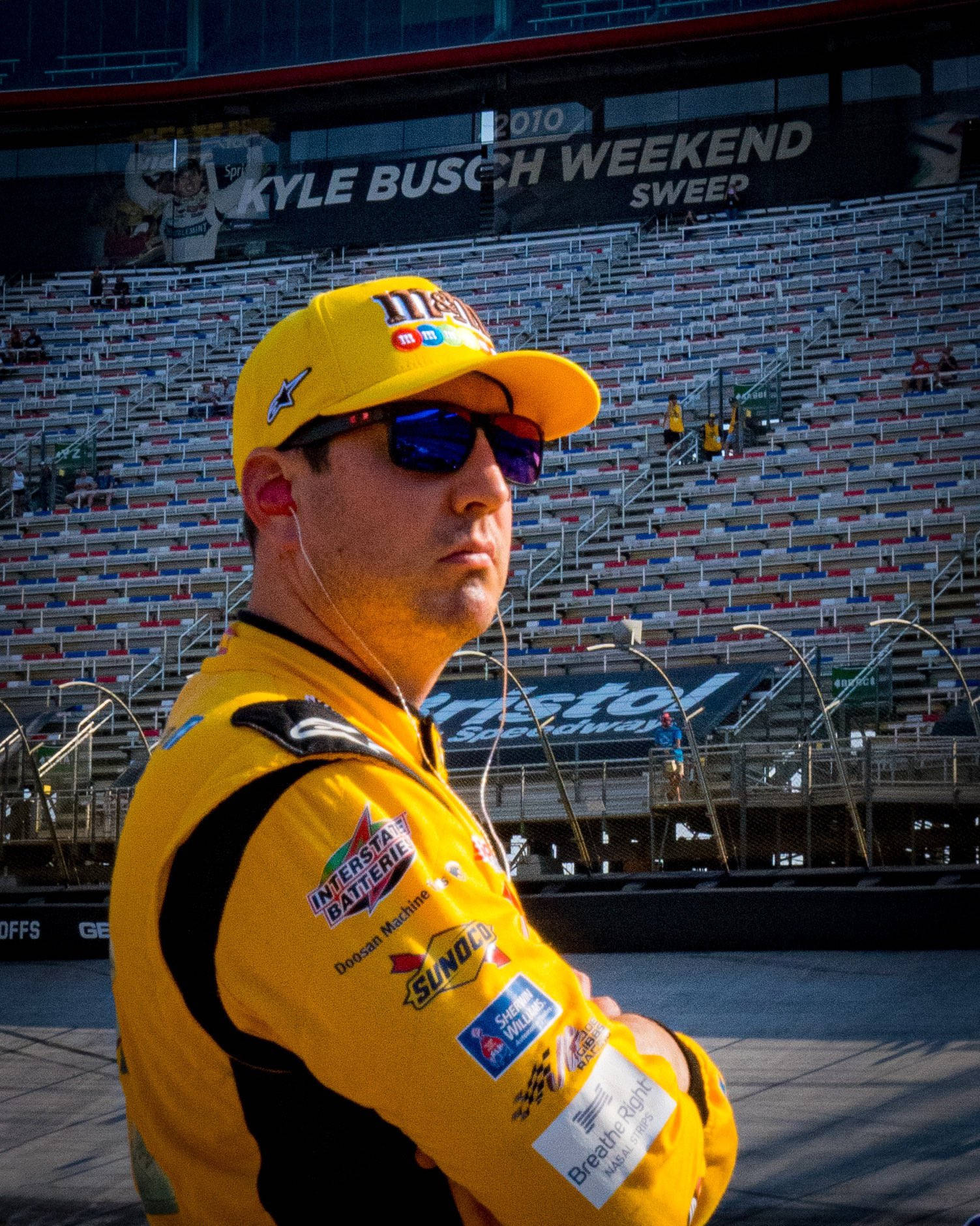 Kyle Busch 在體育場 壁紙 — NASCAR車手凱爾·布施身穿黃色賽車裝備，在布里斯托爾賽道明亮的陽光下自信地交叉雙臂站立。