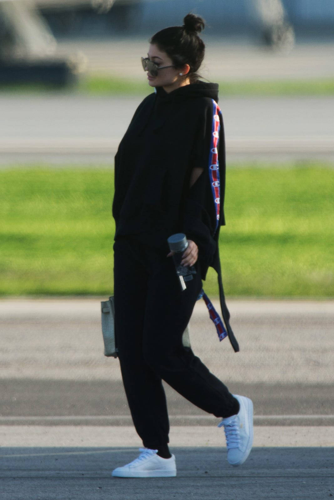 穿著黑色連帽衫和白色運動鞋的女性手持水樽走在機場停機坪上。