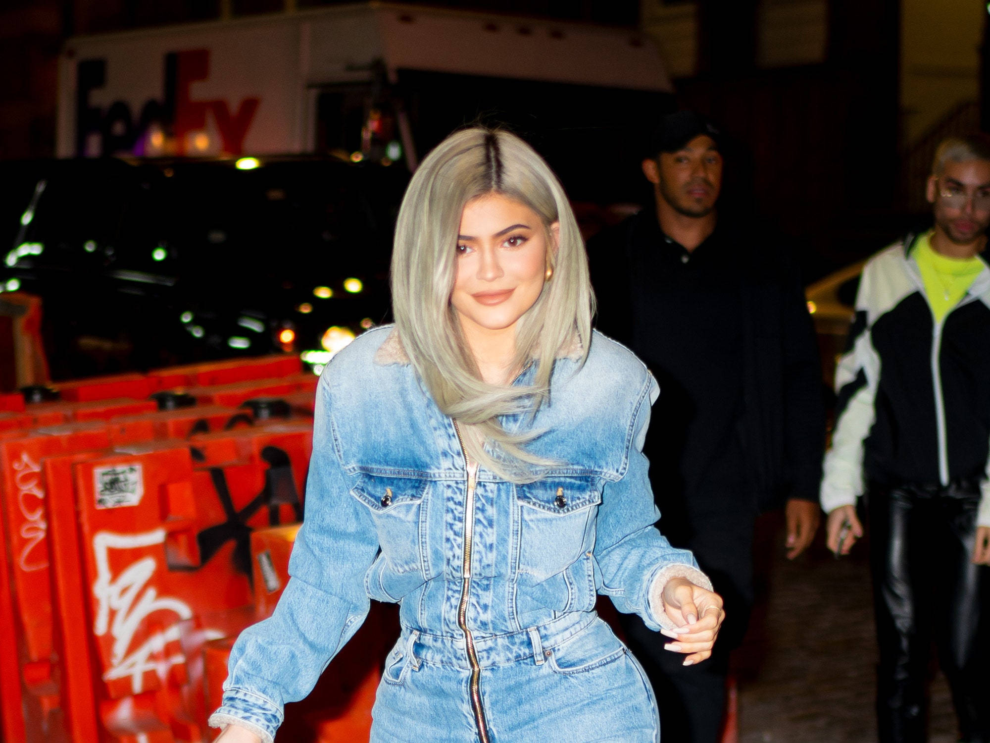 IRO Kylie Jenner 壁紙 — Kylie Jenner 身穿牛仔連身褲，在城市燈光下自信地行走，身後有模糊的行人和一輛FedEx貨車。