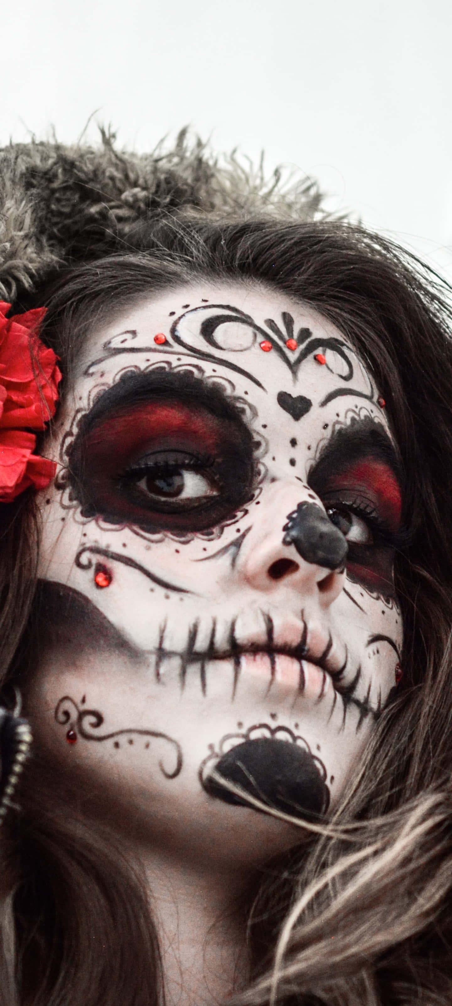 臉上繪有La Calavera彩繪的墨西哥女性肖像 壁紙 — 一位女士的近距離肖像，臉上帶有精緻的糖骷髏妝容，配有紅色點綴和頭髮上的紅花。