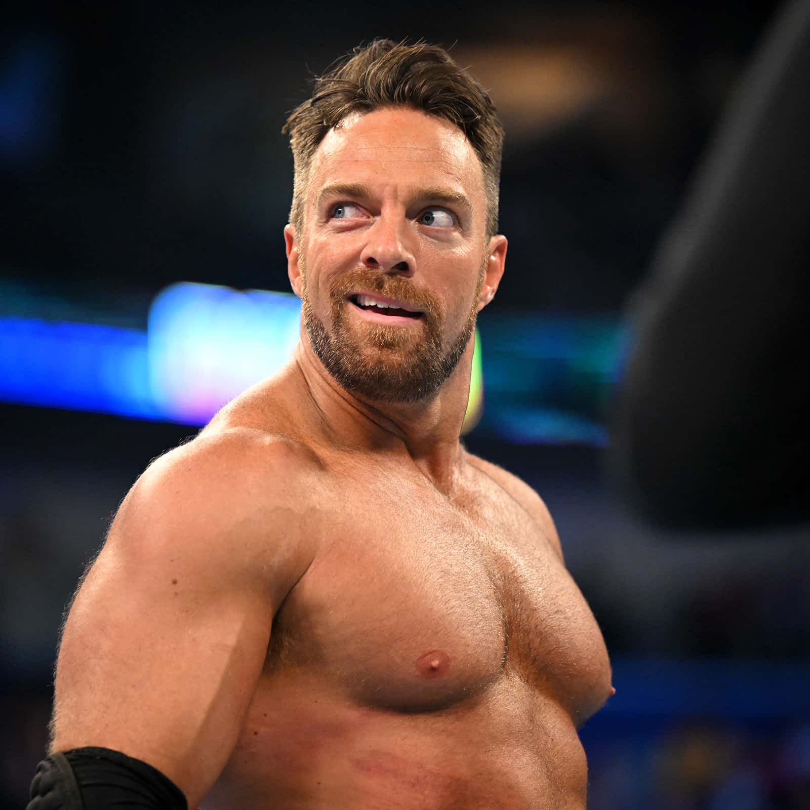 La Knight 在 WWE 活動中自信微笑 壁紙 — 一位肌肉發達、赤裸上身的鬍鬚摔角手在燈光明亮的競技場中自信地向上微笑。