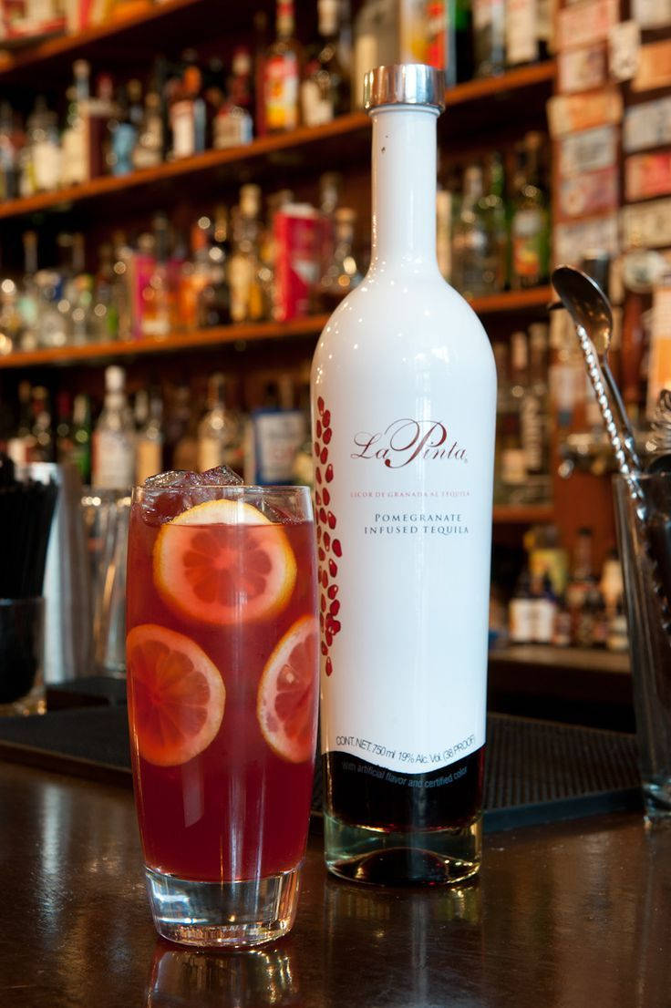 La Pinta石榴利口酒調製誘人柑橘雞尾酒 壁紙 — 一杯充滿活力的石榴注入龍舌蘭雞尾酒，配以柑橘片，放在其酒瓶旁邊，背景是擦亮的吧檯和擺滿酒類的架子。
