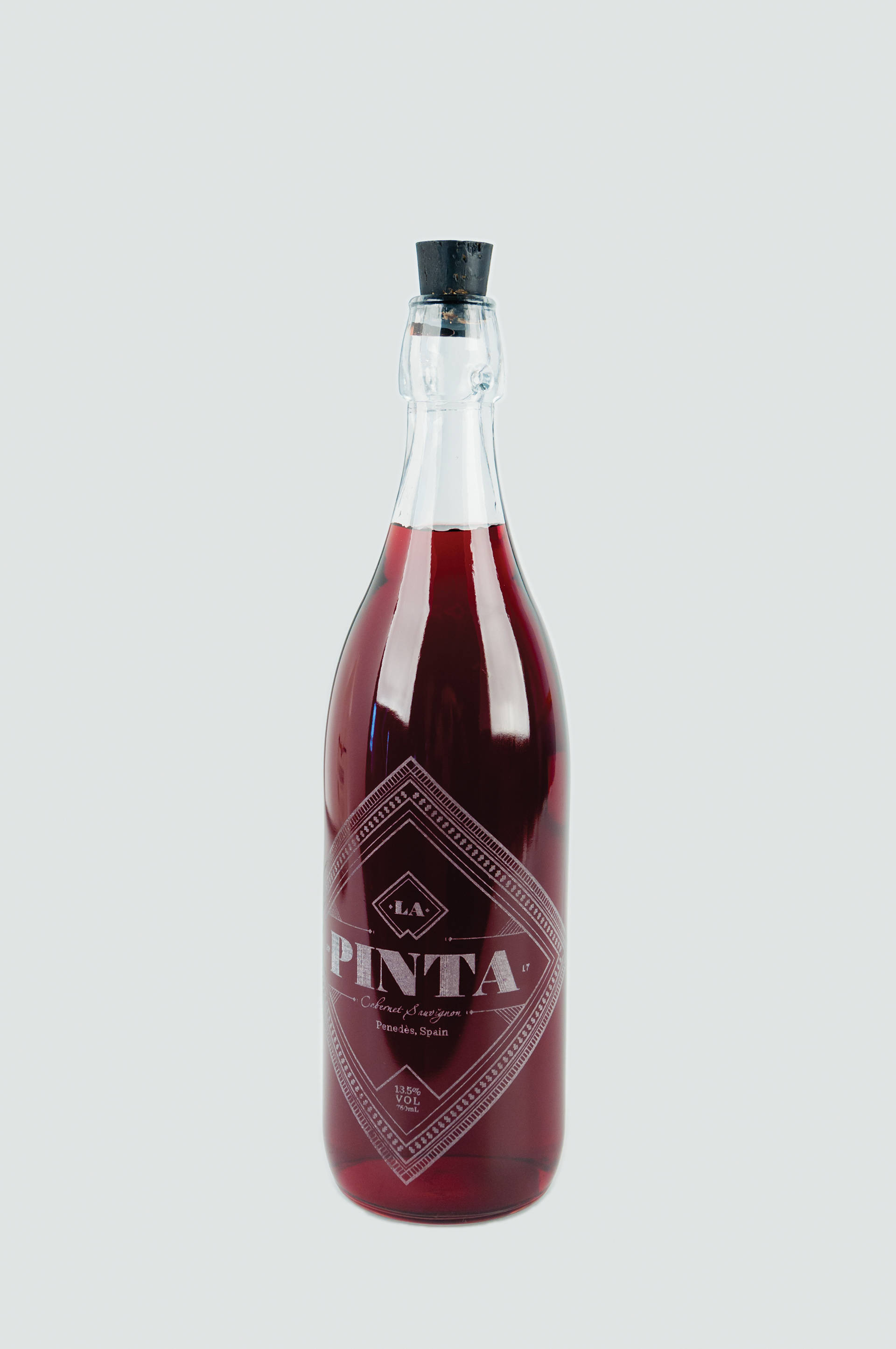 La Pinta石榴利口酒，紅色酒液 壁紙 — 一個高身玻璃瓶，裝滿深紅色液體，配有黑色軟木塞，置於淺灰色背景上。