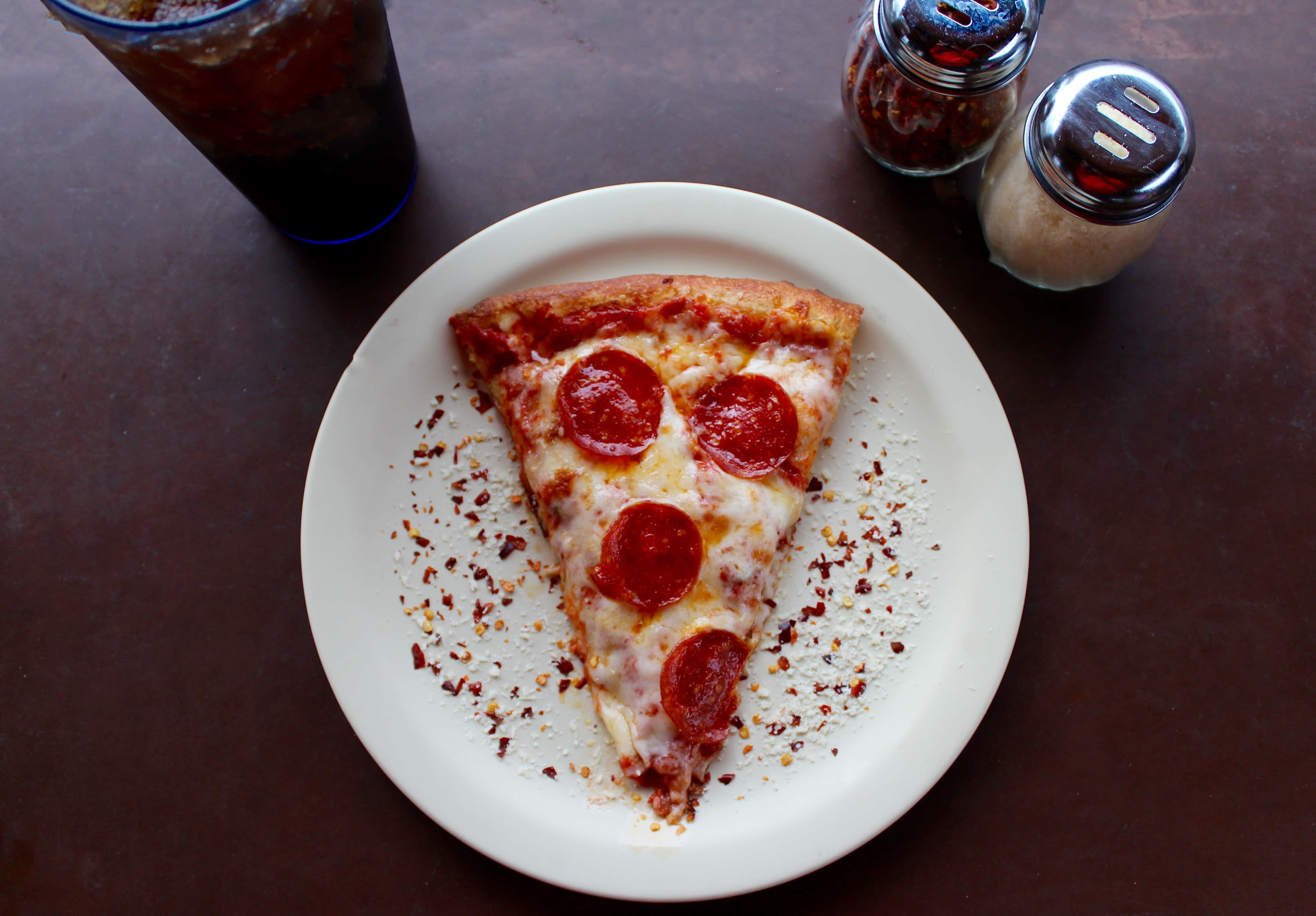 La Pizza Perfetta - 與必勝客一同慶祝 壁紙 — 一片意大利辣香腸比薩放在白色碟子上，配上紅辣椒碎和一杯深色飲料。