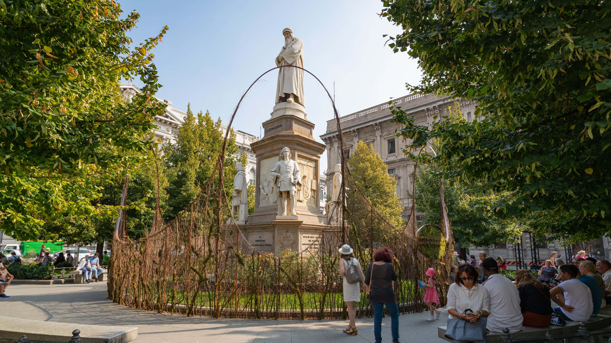 La Scala歌劇院外的雕像 壁紙 — 一個陽光明媚的城市公園場景，人們在歷史雕像周圍放鬆，四周環繞著茂密的綠樹和古典建築。