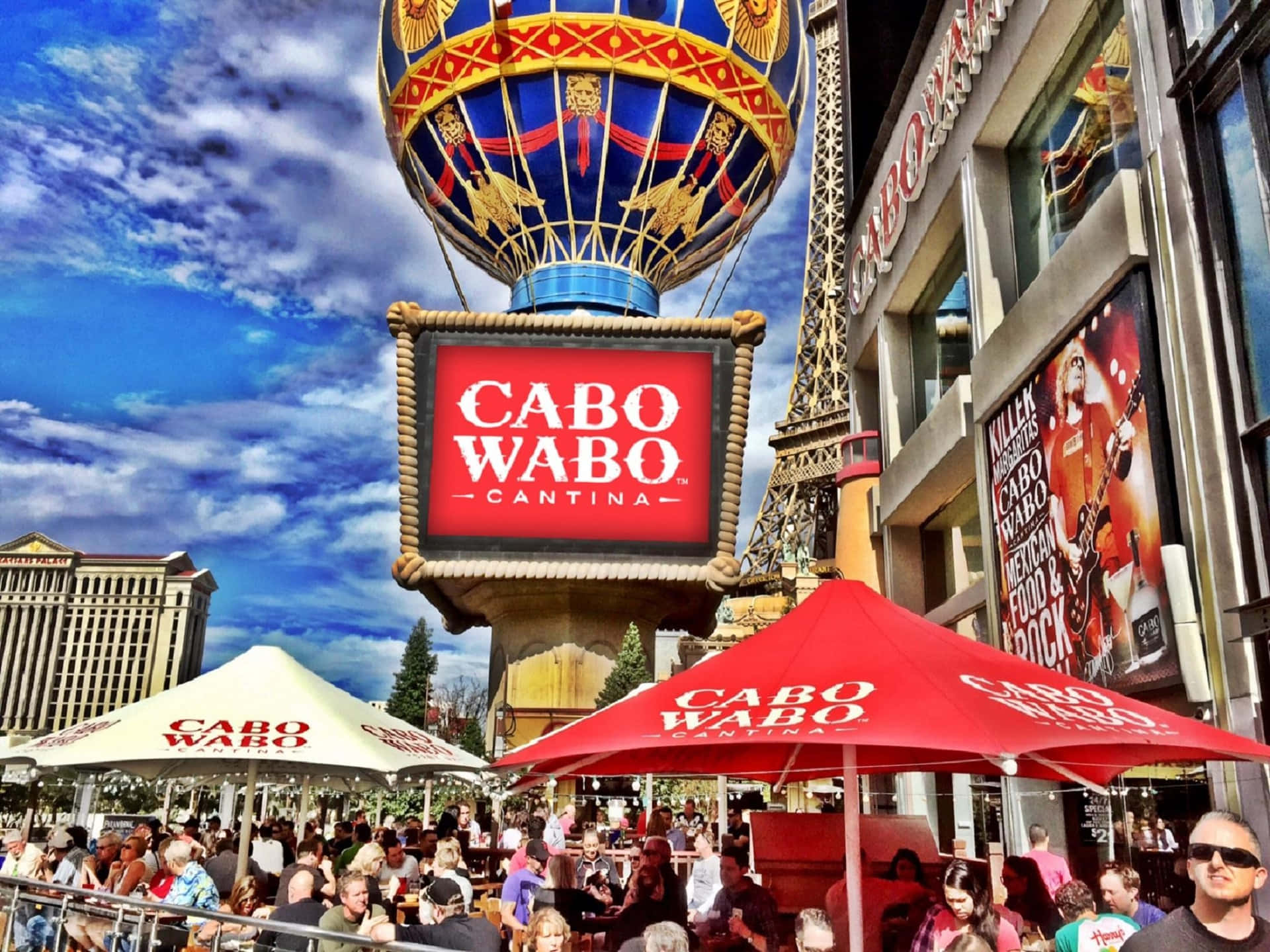  壁紙 — 拉斯維加斯Cabo Wabo Cantina的充滿活力的戶外庭院，設有紅色遮陽傘和一個巨大的熱氣球標誌，背景是明亮的藍天。