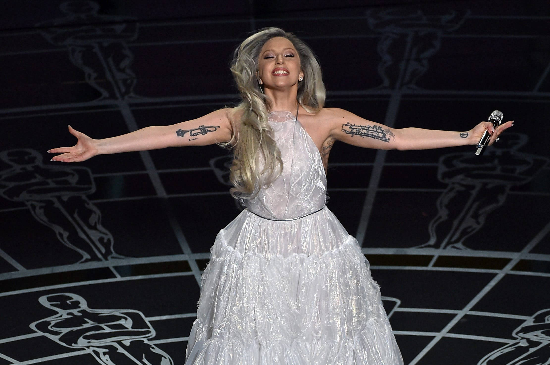 Lady Gaga 2015 奧斯卡頒獎典禮 壁紙 — Lady Gaga 在舞台上表演，身穿閃閃發光的白色長袍，雙臂伸展，手持麥克風。