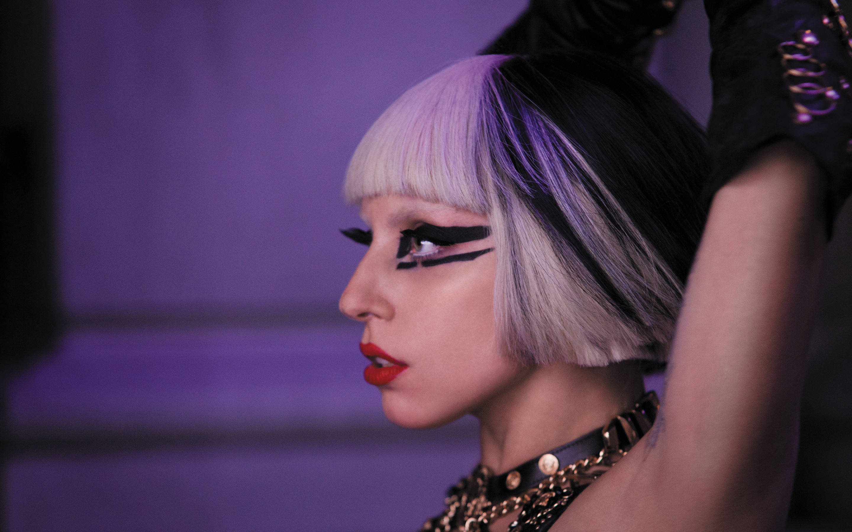 Lady Gaga Edge Of Glory 壁紙 — 一個擁有粗黑翼形眼線和紫色燈光下銀色頭髮的人，在黑暗華麗的場景中擺姿勢。