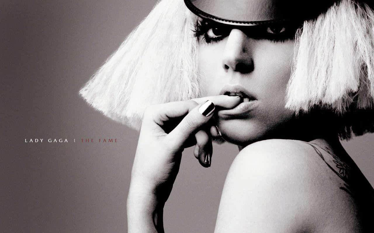 Lady Gaga 桌布 壁紙 — Lady Gaga 以單色調呈現，戴著白色鮑伯頭假髮和深色太陽眼鏡，手指靠近嘴唇，肩膀上的刺青清晰可見。