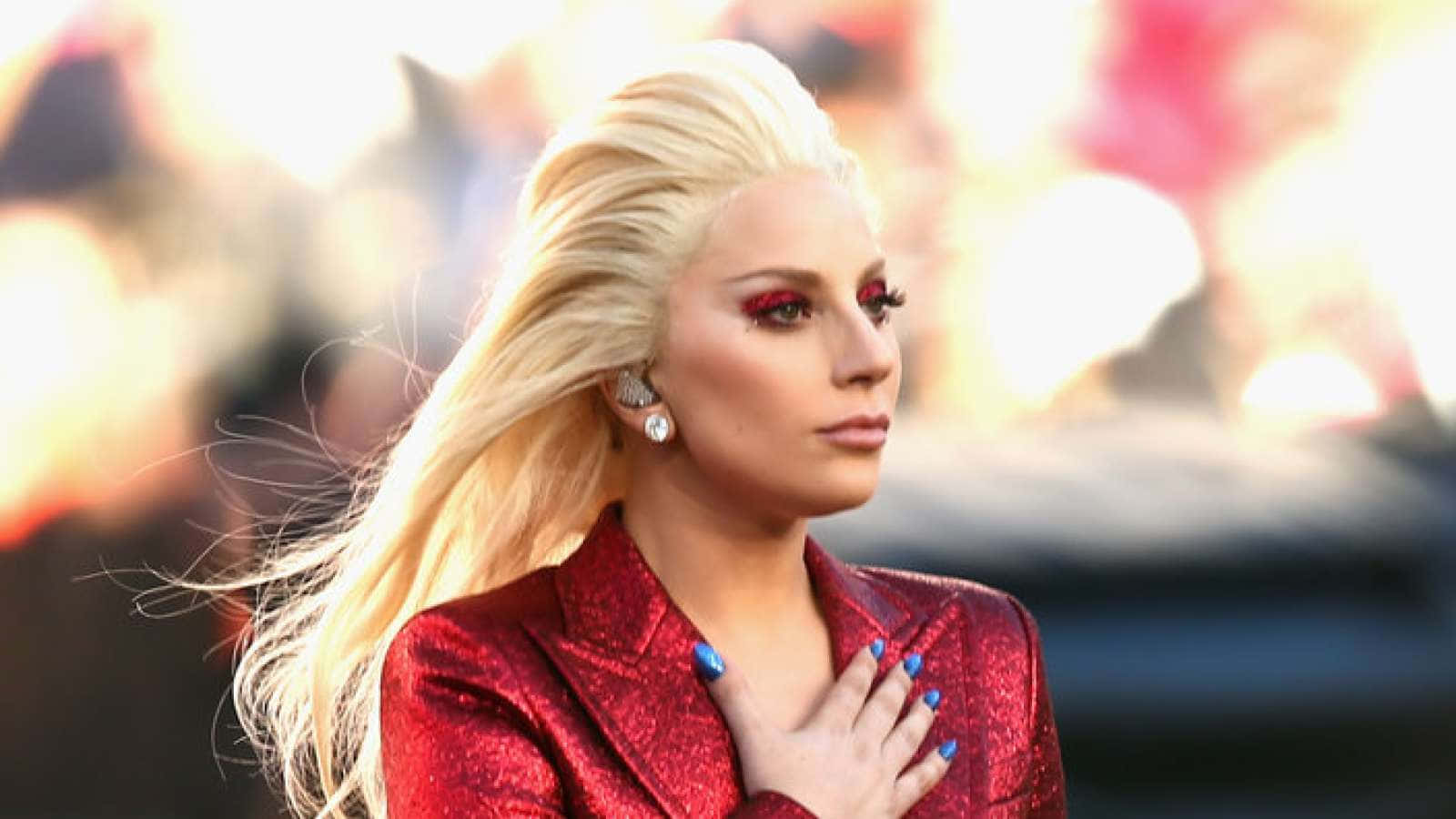 Lady Gaga 桌布 壁紙 — Lady Gaga 身穿閃亮紅色外套，手放胸前，金髮飄逸，大膽紅色眼妝，背景模糊火焰。