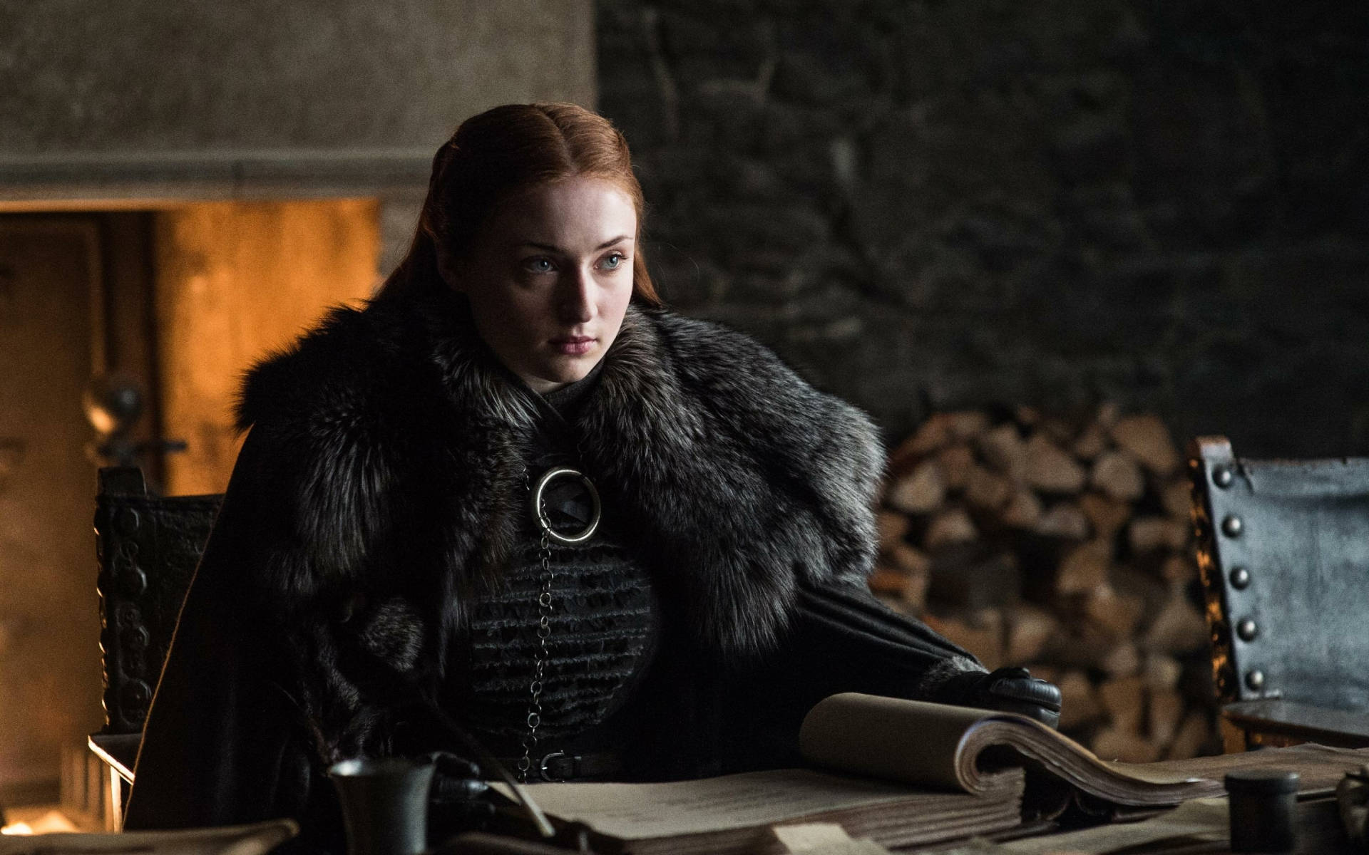 聰明的Lady Sansa Stark 壁紙 — 一位身穿毛皮襯裡黑色斗篷的女性坐在一張木桌前，身後是壁爐的昏暗石室中。