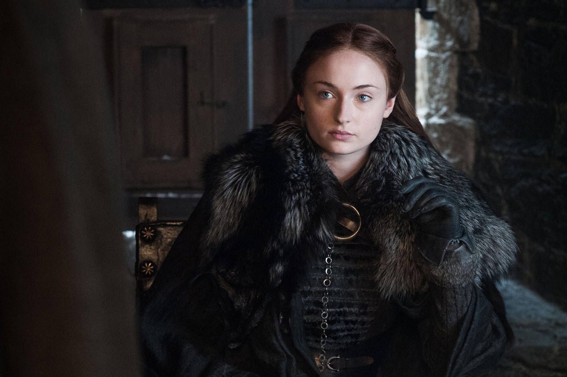 Lady Sansa Stark 壁紙 — 一位身穿深色毛皮襯裡斗篷的女性，端莊地坐在石牆房間中，擁有深邃的藍色眼睛和嚴肅的表情。