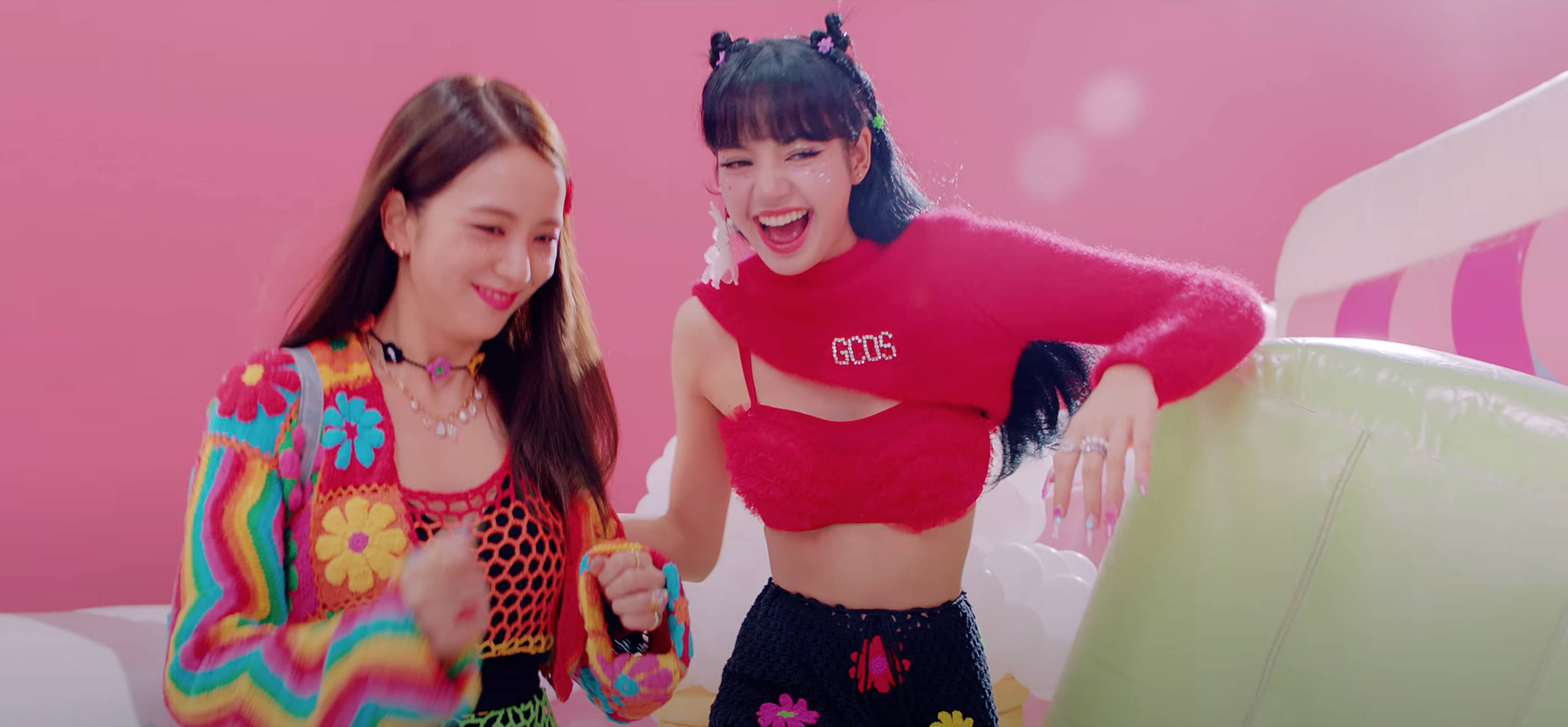 Lalisa Manoban 與 BLACKPINK Jisoo 壁紙 — 兩位快樂的女性穿著鮮豔多彩的服裝，在明亮的粉紅色背景和充氣道具前一起大笑。