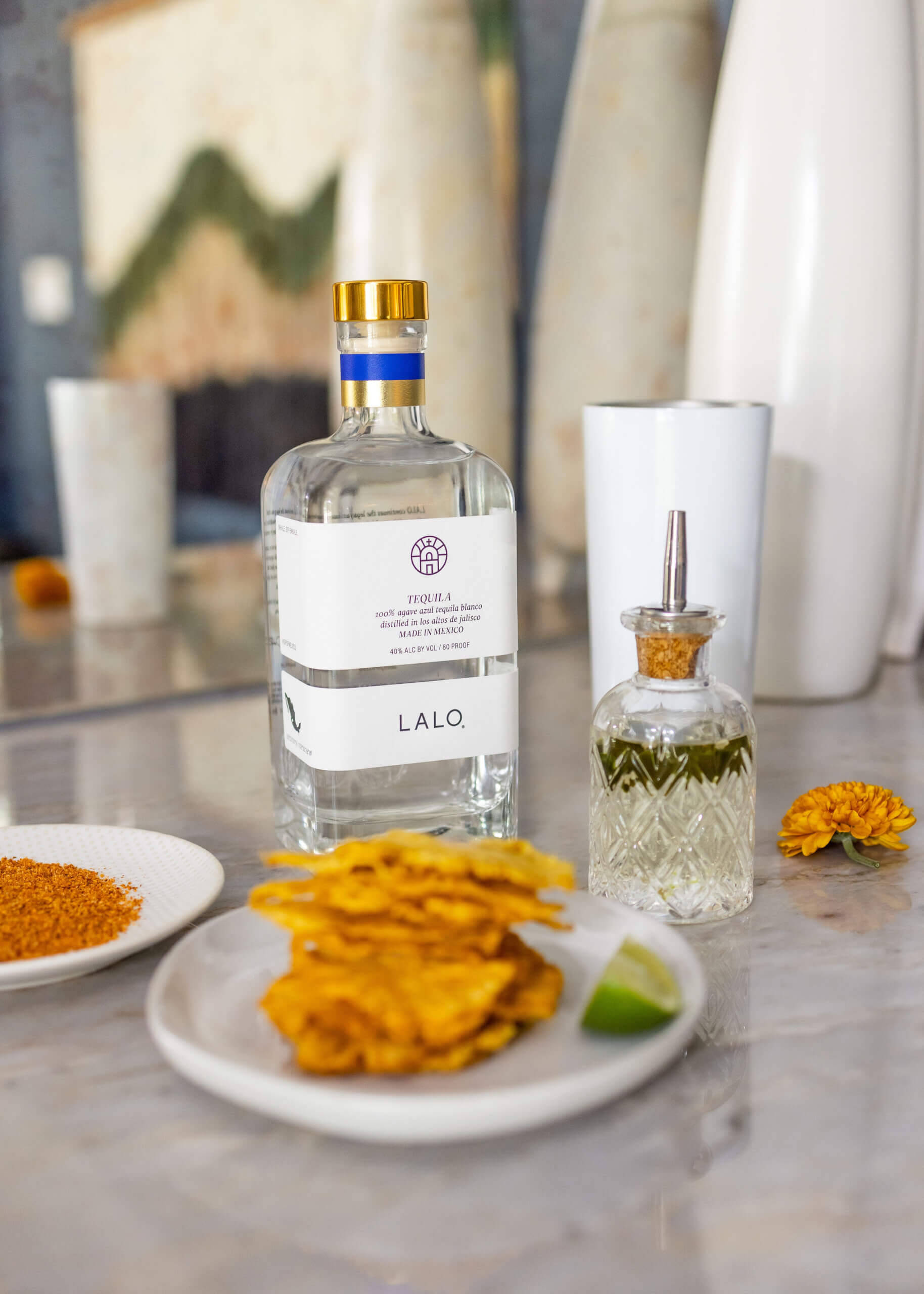 品味Lalo龍舌蘭酒搭配酥脆小食 壁紙 — 一瓶流線型的Lalo龍舌蘭酒放在大理石檯面上，旁邊有脆薯片、青檸和花卉裝飾。