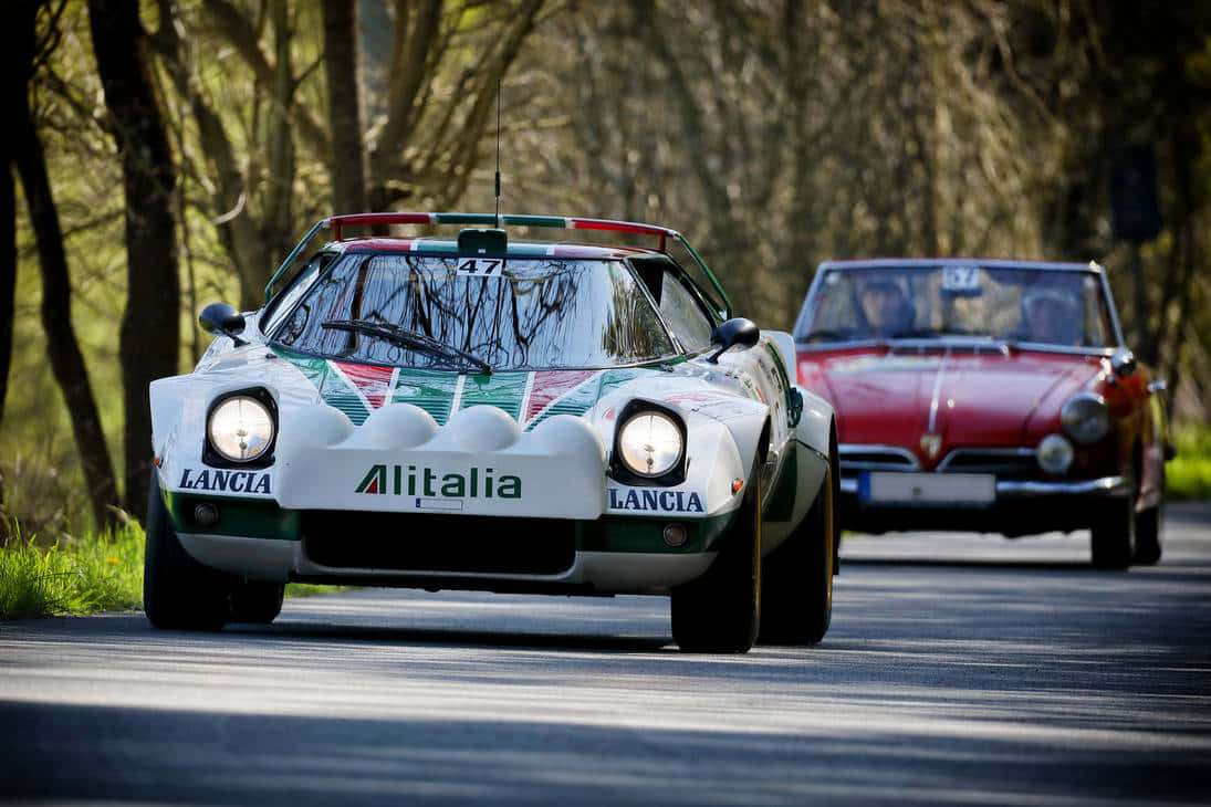 Lancia Stratos Alitalia 賽車 壁紙 — 一架白色 Lancia 賽車帶有 Alitalia 塗裝，在樹木成蔭的道路上飛馳，後面有一輛紅色經典汽車緊隨其後。