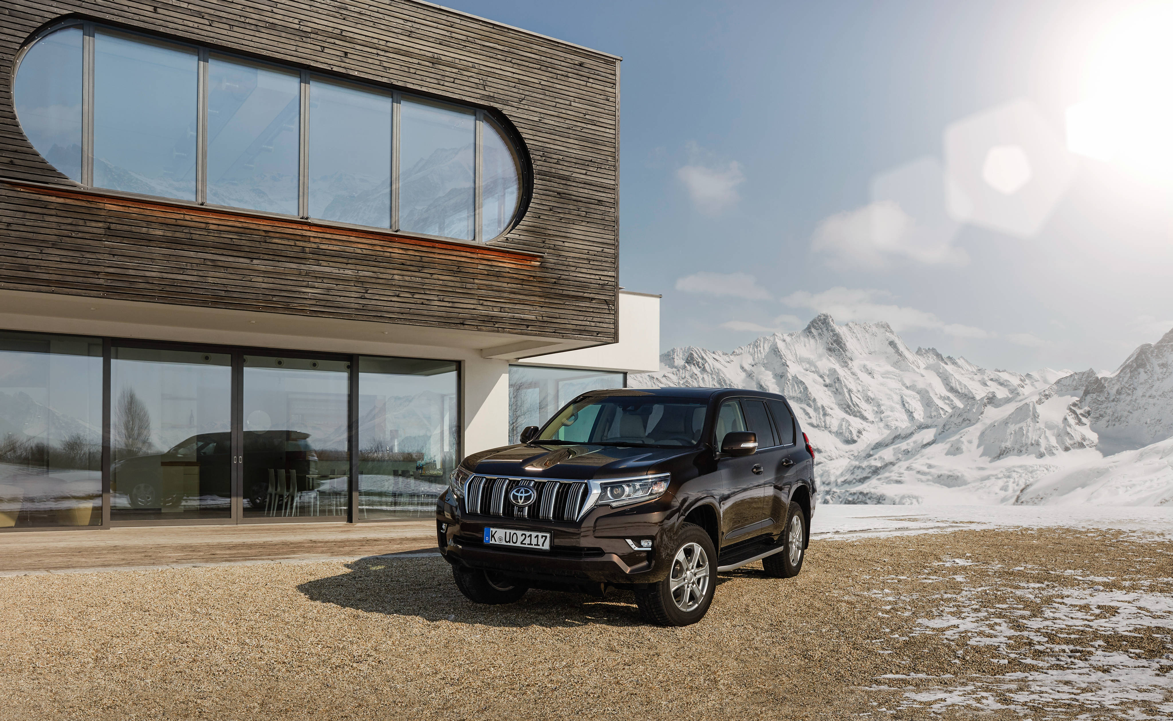 光澤黑漆Land Cruiser Toyota 4K 壁紙 — 一輛時尚的黑色SUV停在一棟現代木屋前，木屋帶有弧形玻璃窗，背景是陽光明媚下白雪覆蓋的山脈。