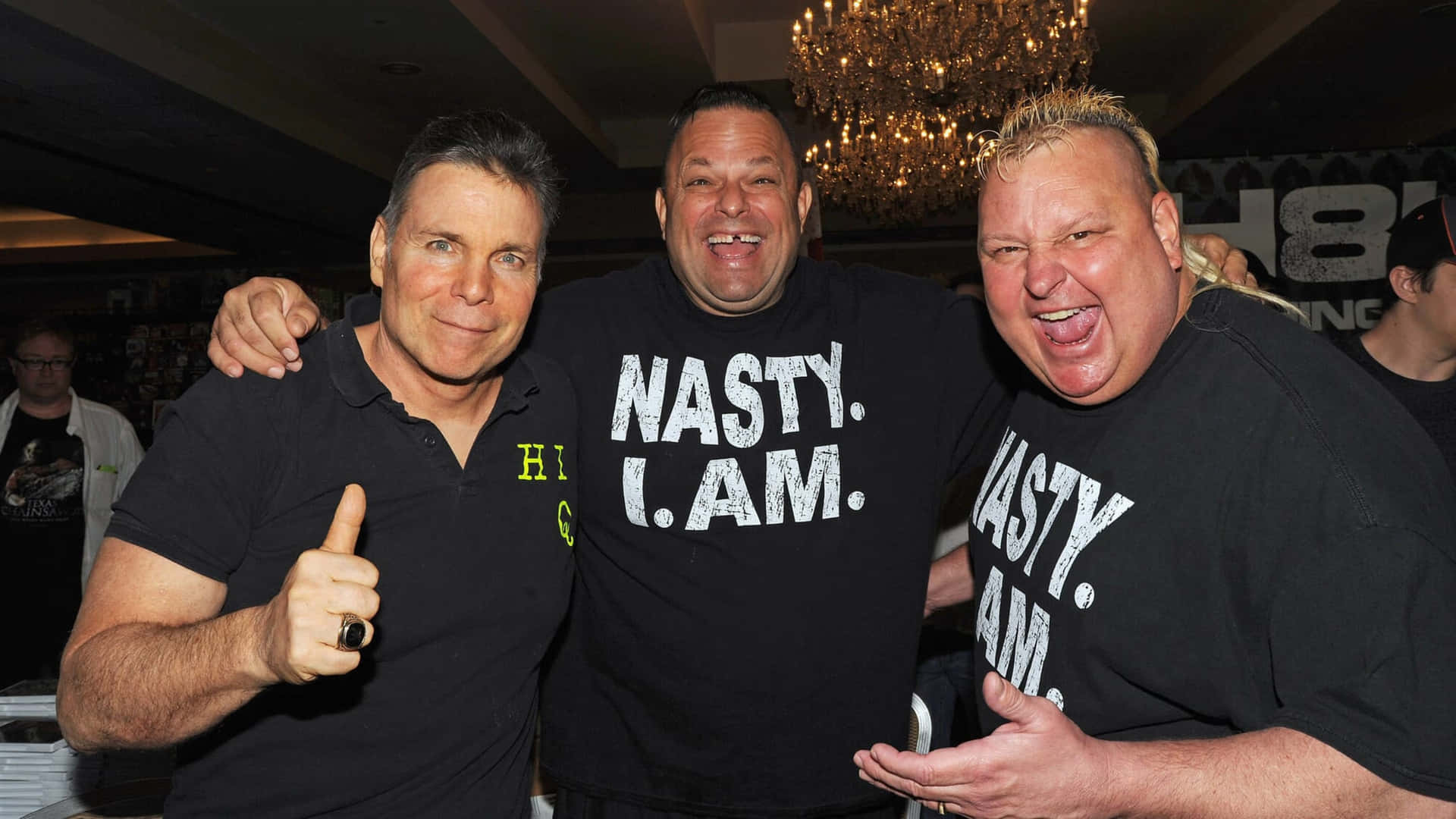 Lanny Poffo 拇指向上 壁紙 — 三名身穿黑色 'NASTY I AM' T恤的男子在昏暗的房間裡，在閃爍的水晶燈下豎起大拇指，愉快地擺姿勢。