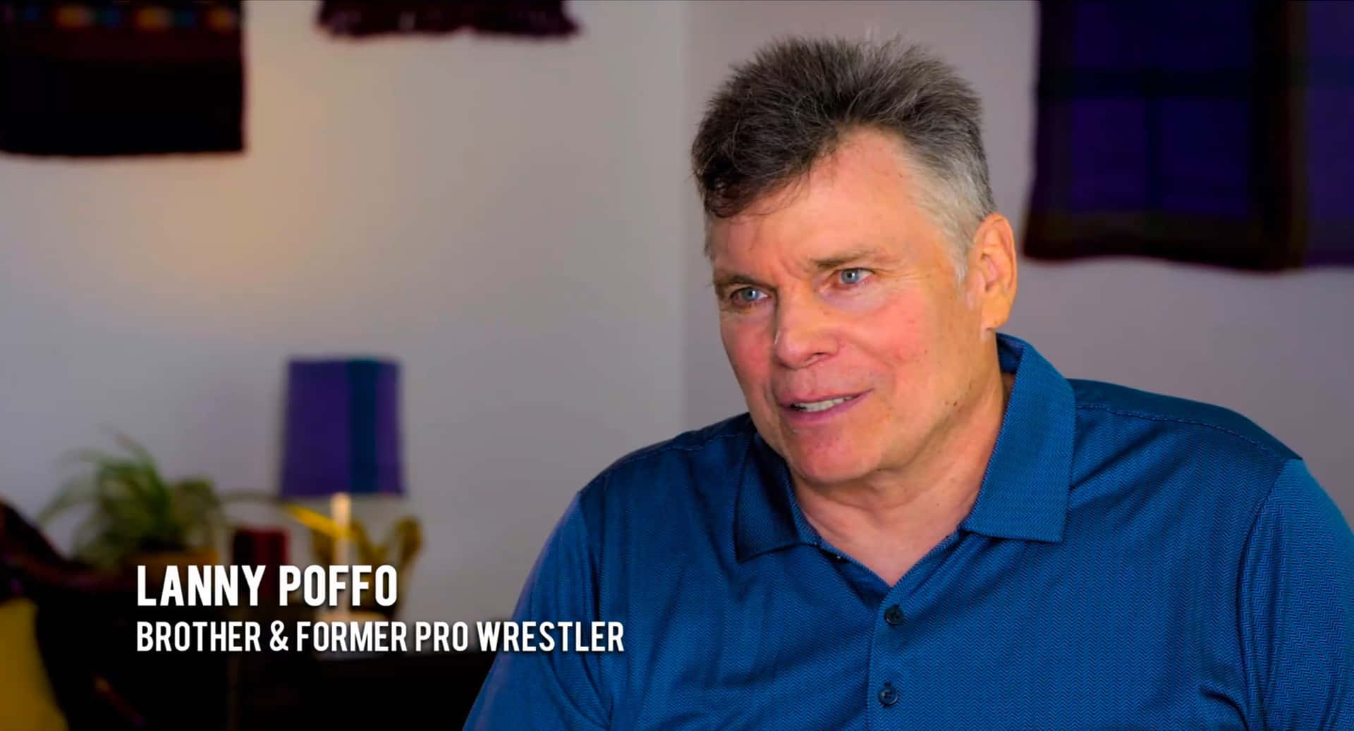 Lanny Poffo 擊掌訪問 壁紙 — 一位灰髮藍眼的中年男子在室內穿著藍色Polo衫微笑。
