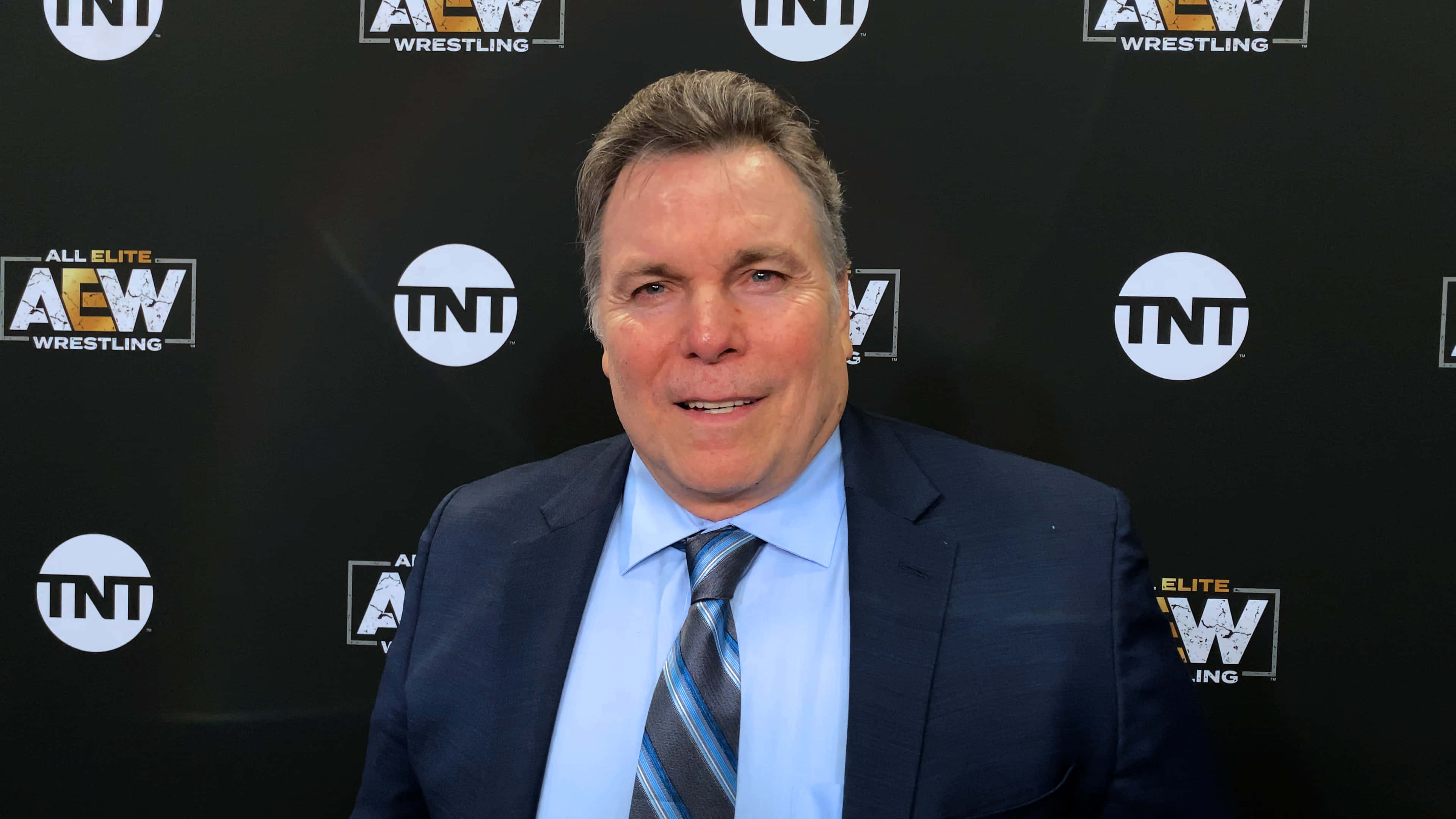 Lanny Poffo 黑西裝藍Polo衫 壁紙 — 一名身穿深色西裝和條紋領帶的男子在帶有AEW和TNT標誌的黑色背景下對著鏡頭微笑。