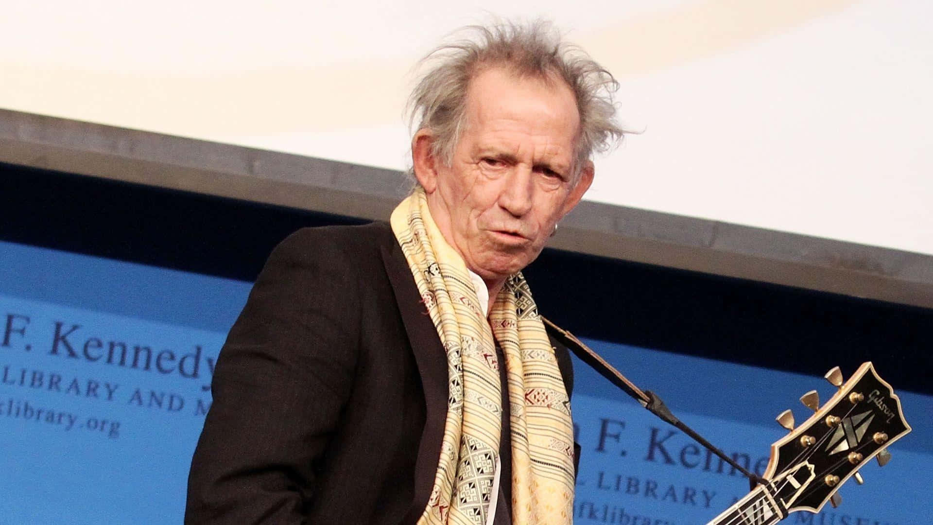  壁紙 — 一位頭髮灰白蓬鬆的老年男子，戴著圖案圍巾和黑色外套，在背景為 'F Kennedy Library' 的講台上拿著結他。