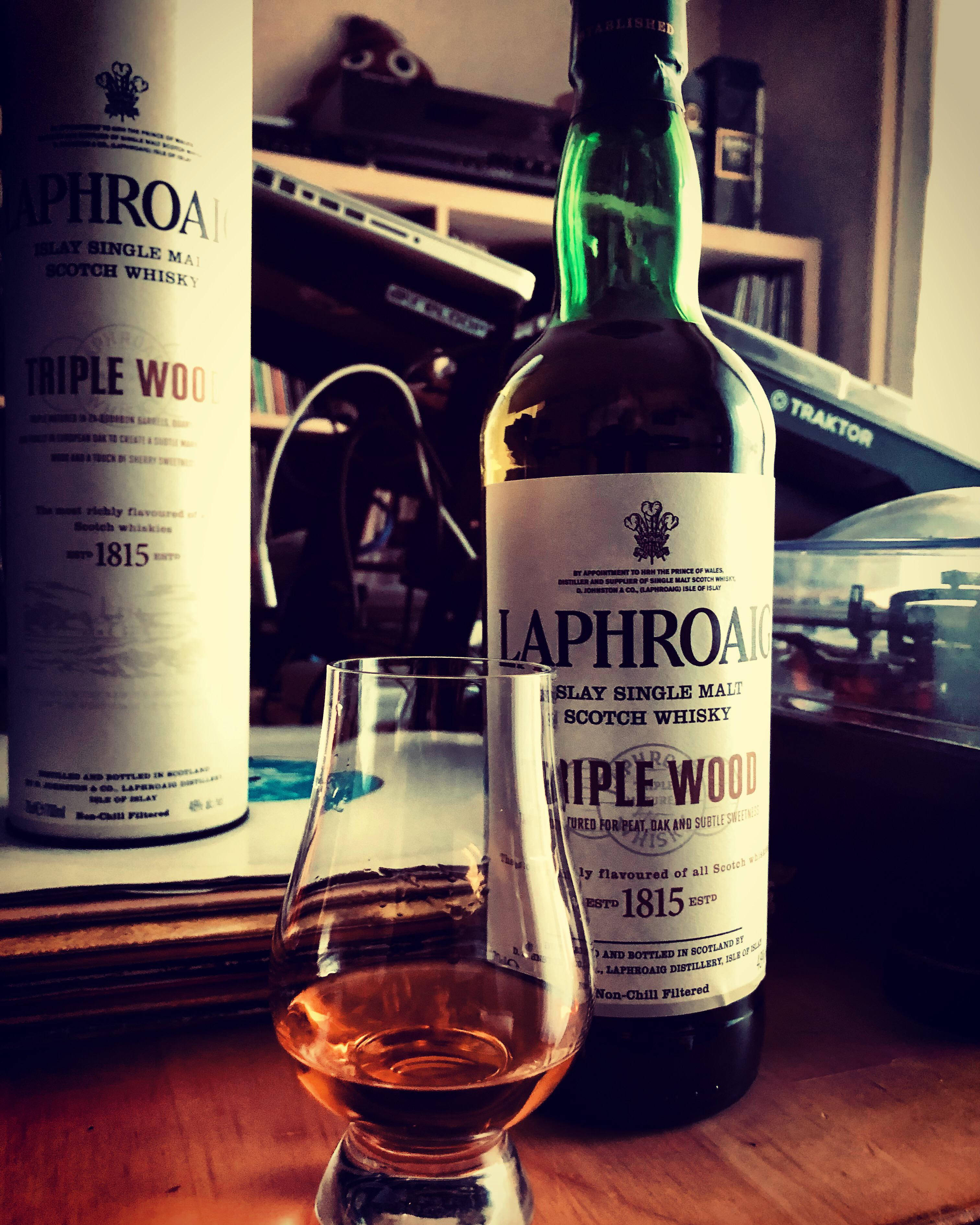 Laphroaig Triple Wood威士忌 壁紙 — 兩瓶Laphroaig Triple Wood威士忌和一個品酒杯放在木桌上，房間光線溫暖，背景有音響設備。