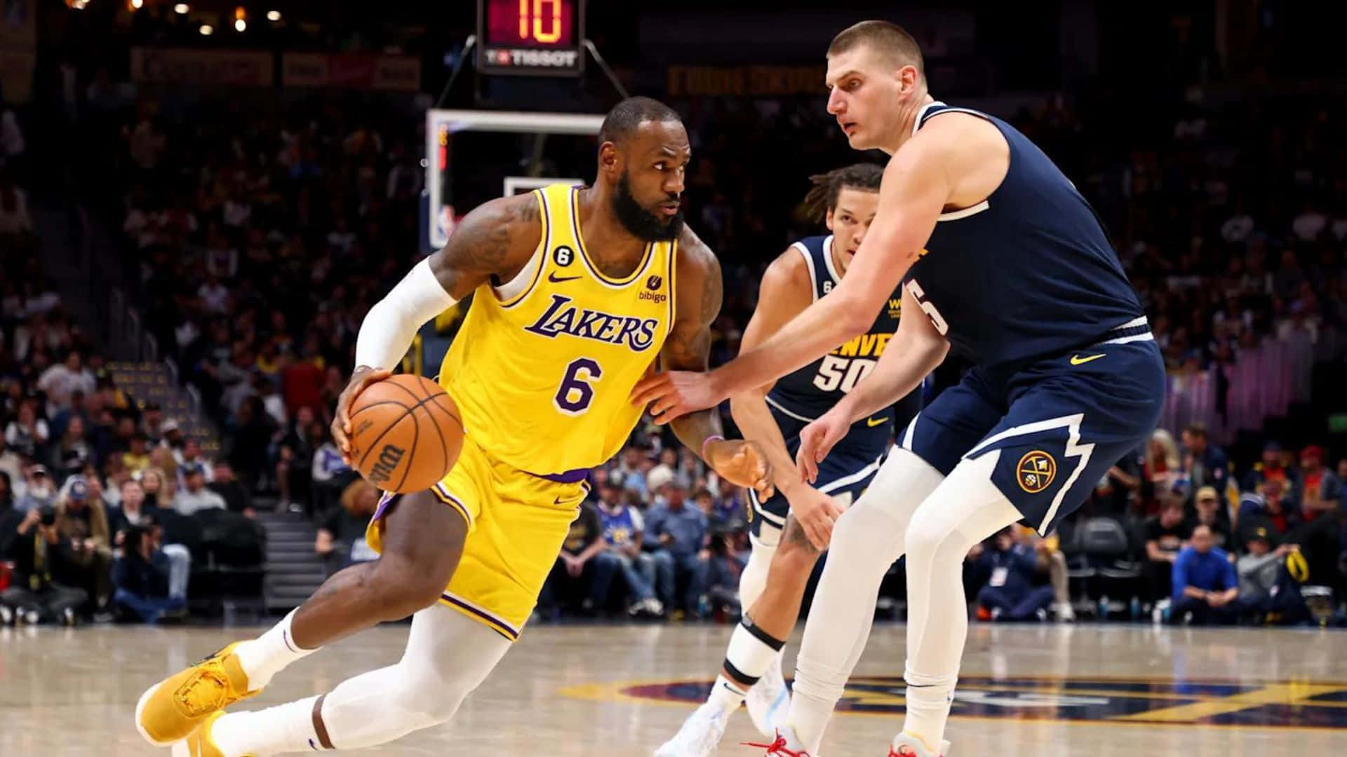 NBA Game 桌布 壁紙 — 勒邦占士身穿黃色湖人球衣，在場館燈光下於一場激烈的NBA比賽中運球越過丹佛金塊防守球員。