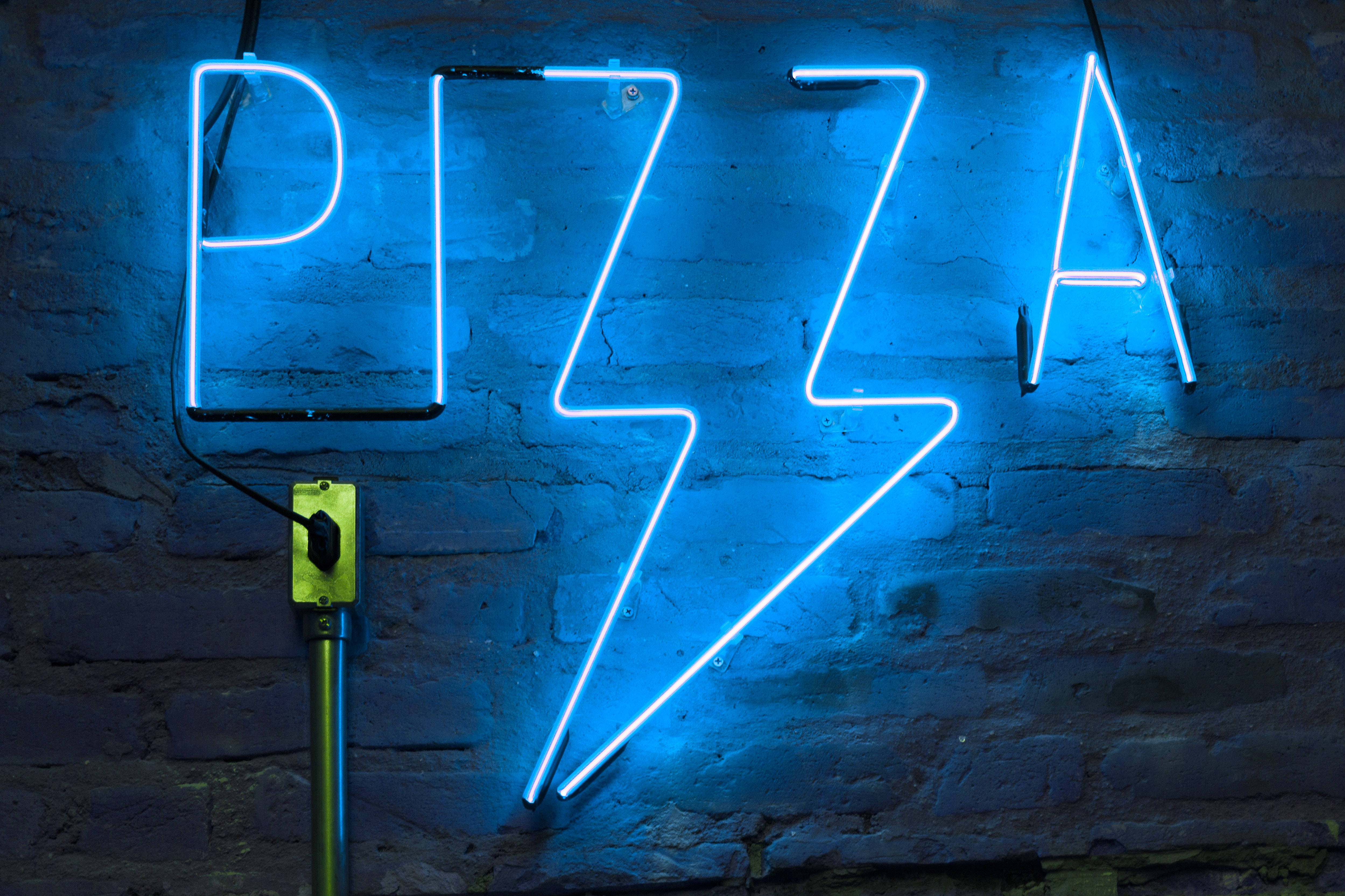 美學藍色披薩LED燈 壁紙 — 一個發光的藍色霓虹燈牌，拼出 'PIZZA' 字樣，帶有閃電形狀，安裝在深色磚牆上。