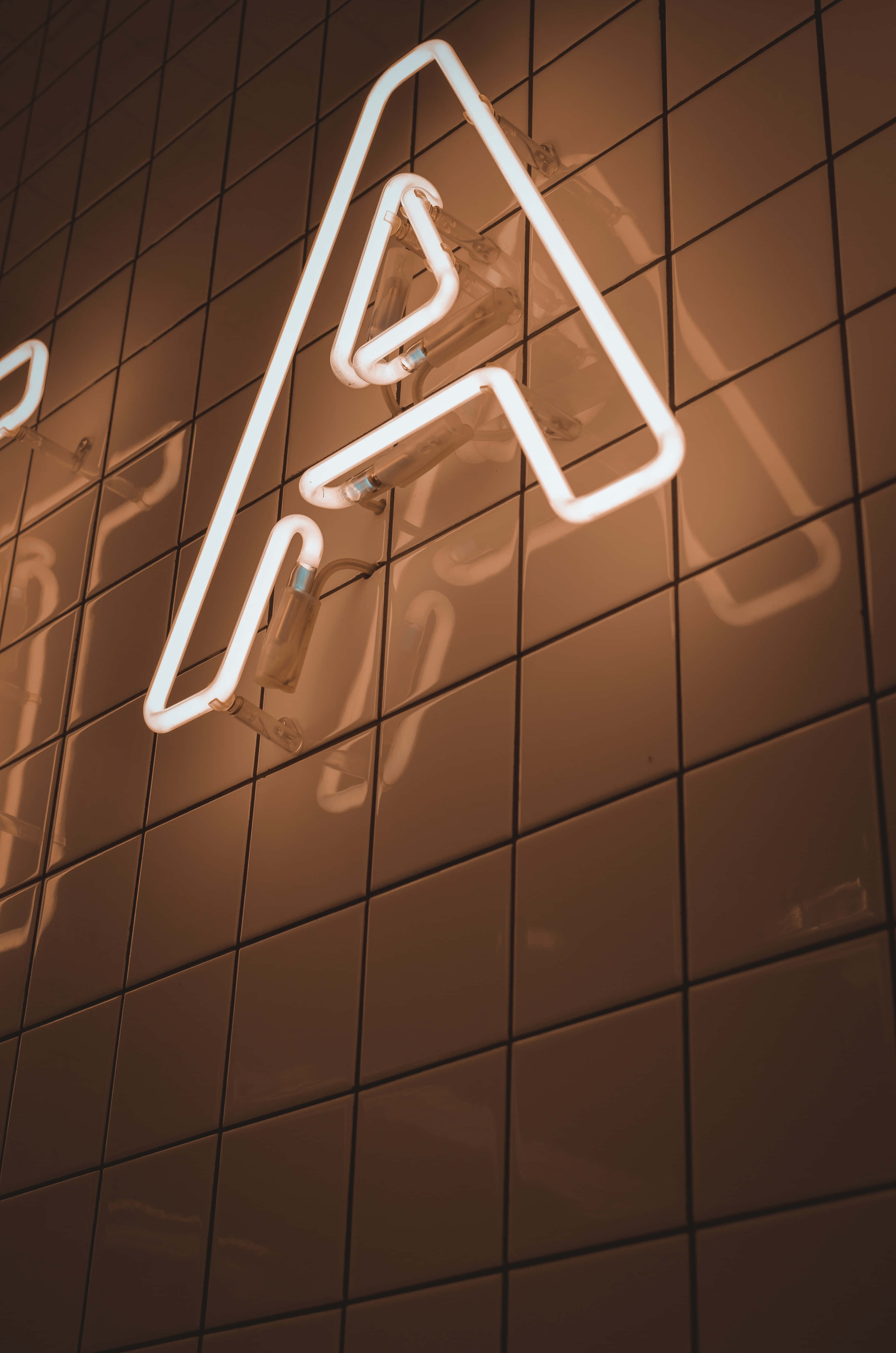 LED白色字母A背景 壁紙 — 一個發光的白色霓虹字母A安裝在深色瓷磚牆上，散發出溫暖的環境光。