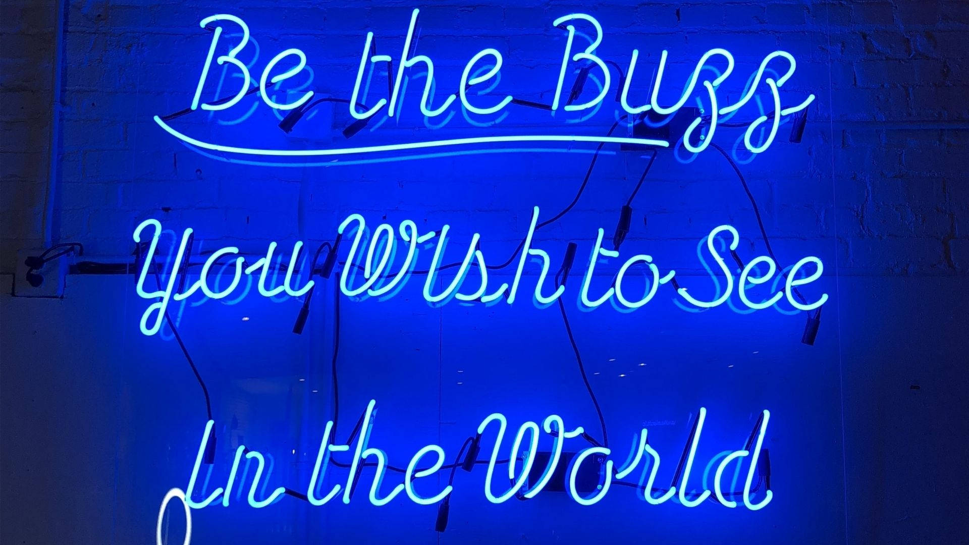 LED霓虹藍標誌 壁紙 — 一個發光的藍色霓虹燈牌，上面寫著 'Be the Buzz You Wish to See In the World'，背景是深色磚牆。