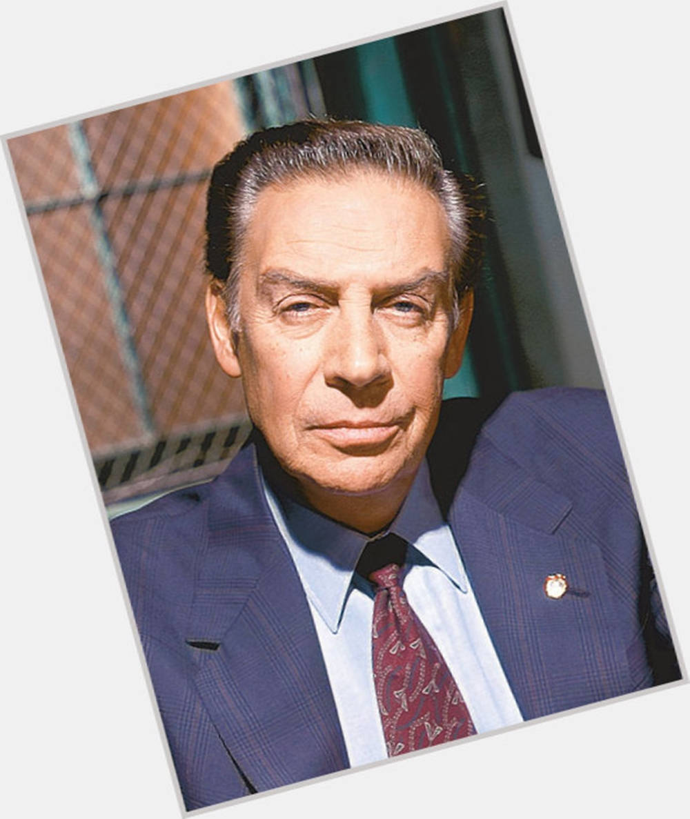 Legendary American Actor Jerry Orbach Portraying Detective Lennie Briscoe 壁紙 — 一位穿著藍色細條紋西裝和圖案領帶的衣著講究男士，在模糊的城市背景下專注地凝視著相機。
