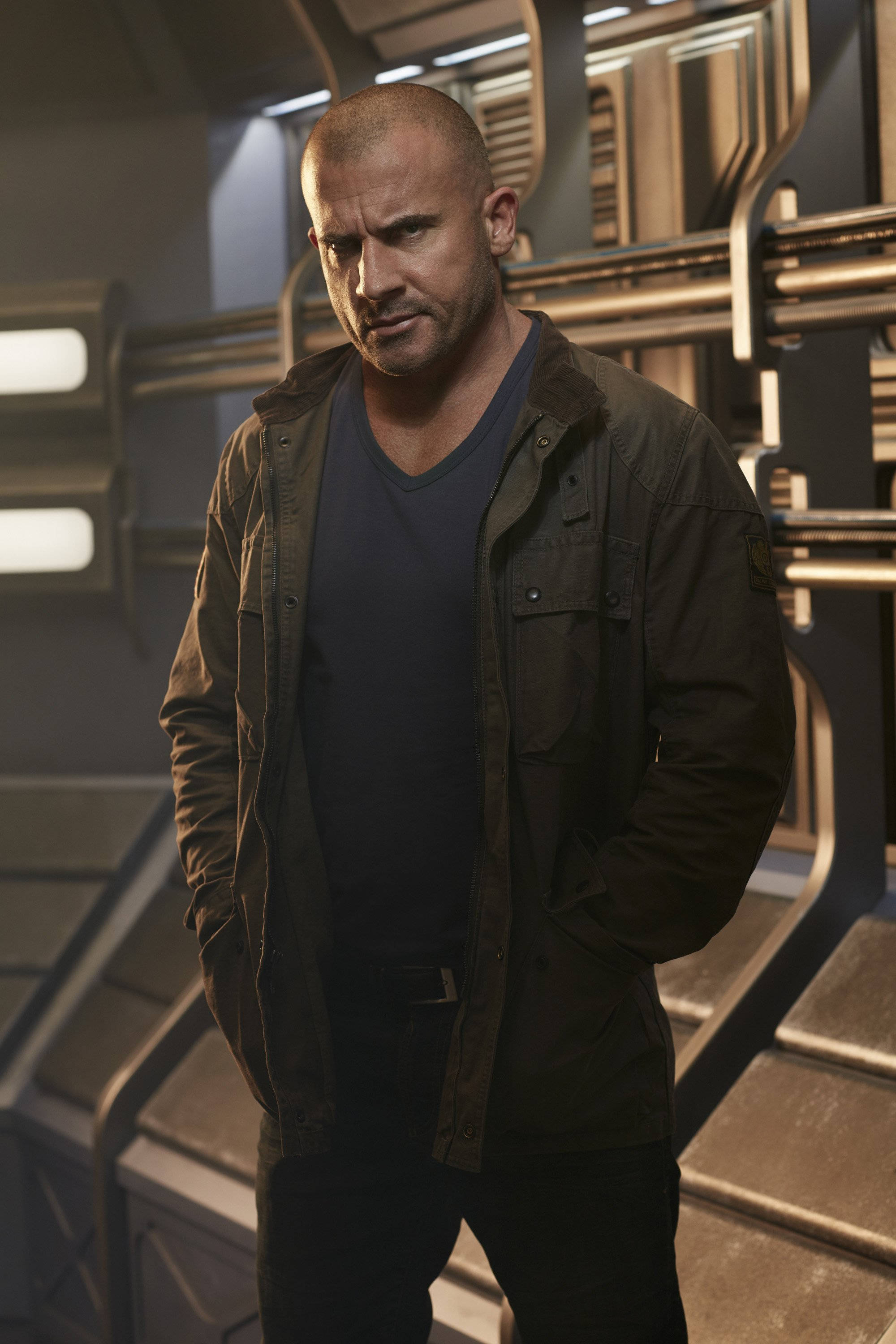 傳奇明日 LEGENDS OF TOMORROW 米克·羅里 壁紙 — 一個光頭男子穿著深色外套，雙手插袋，站在未來風格的金屬太空船走廊內。