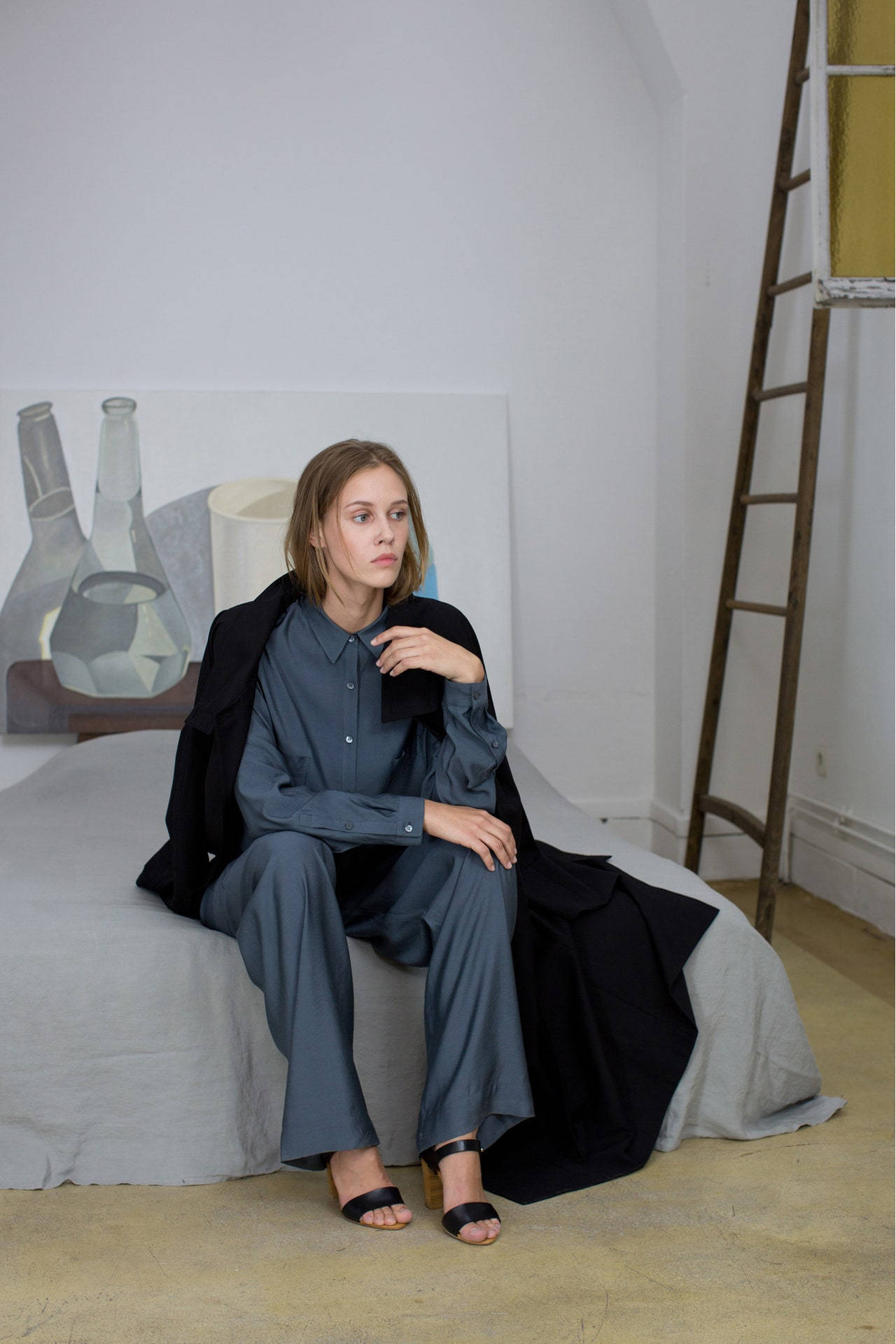 身穿藍色服裝的女性坐在帶有梯子和藝術品的極簡白色房間的床上。