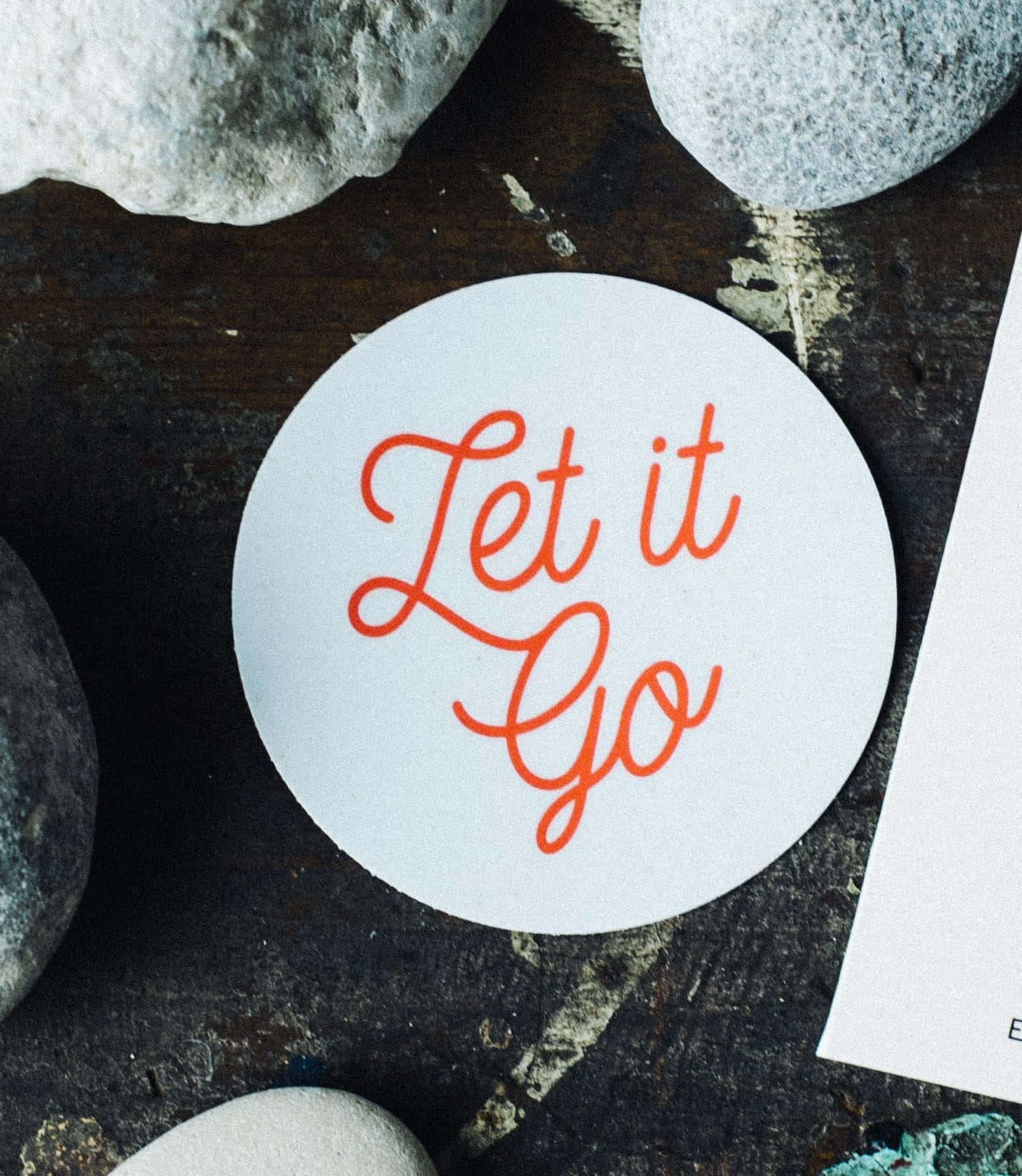 Let It Go 環繞岩石 壁紙 — 一個白色圓形貼紙，上面有橙色草書文字寫著「Let it Go」，放在深色紋理表面的灰色河石之間。