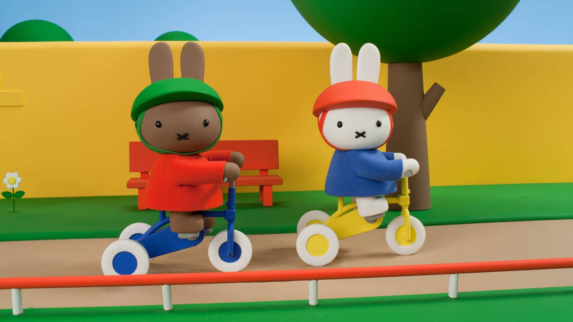 Miffy與Melanie騎自行車 壁紙 — 兩個黏土動畫兔子角色喺綠色樹下嘅明亮公園小徑上踩住色彩繽紛嘅三輪車。