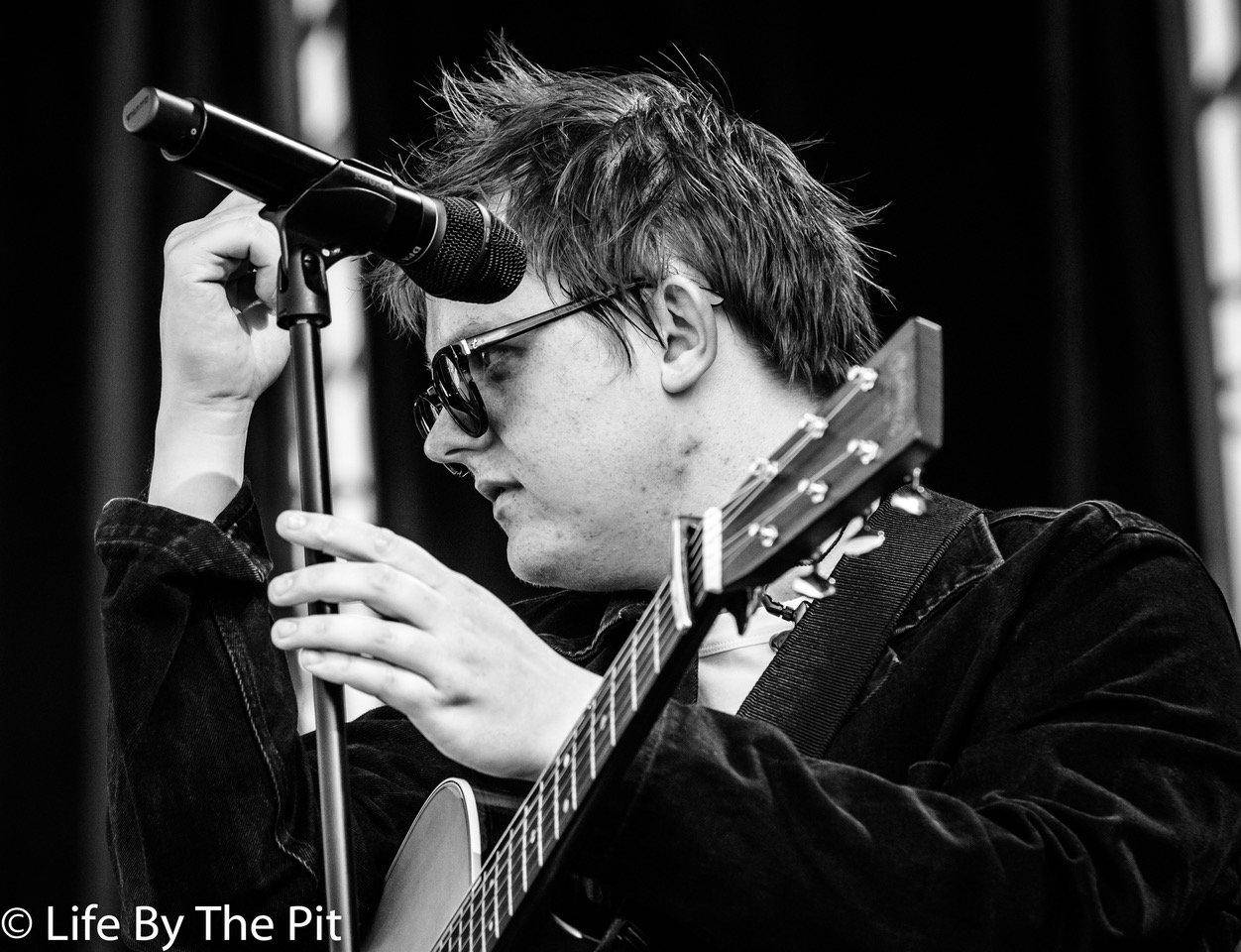 Lewis Capaldi 2019 Mad Cool 音樂節黑白攝影 壁紙 — 一位戴著太陽眼鏡的音樂家在黑白表演照中調整麥克風，同時彈奏木結他。
