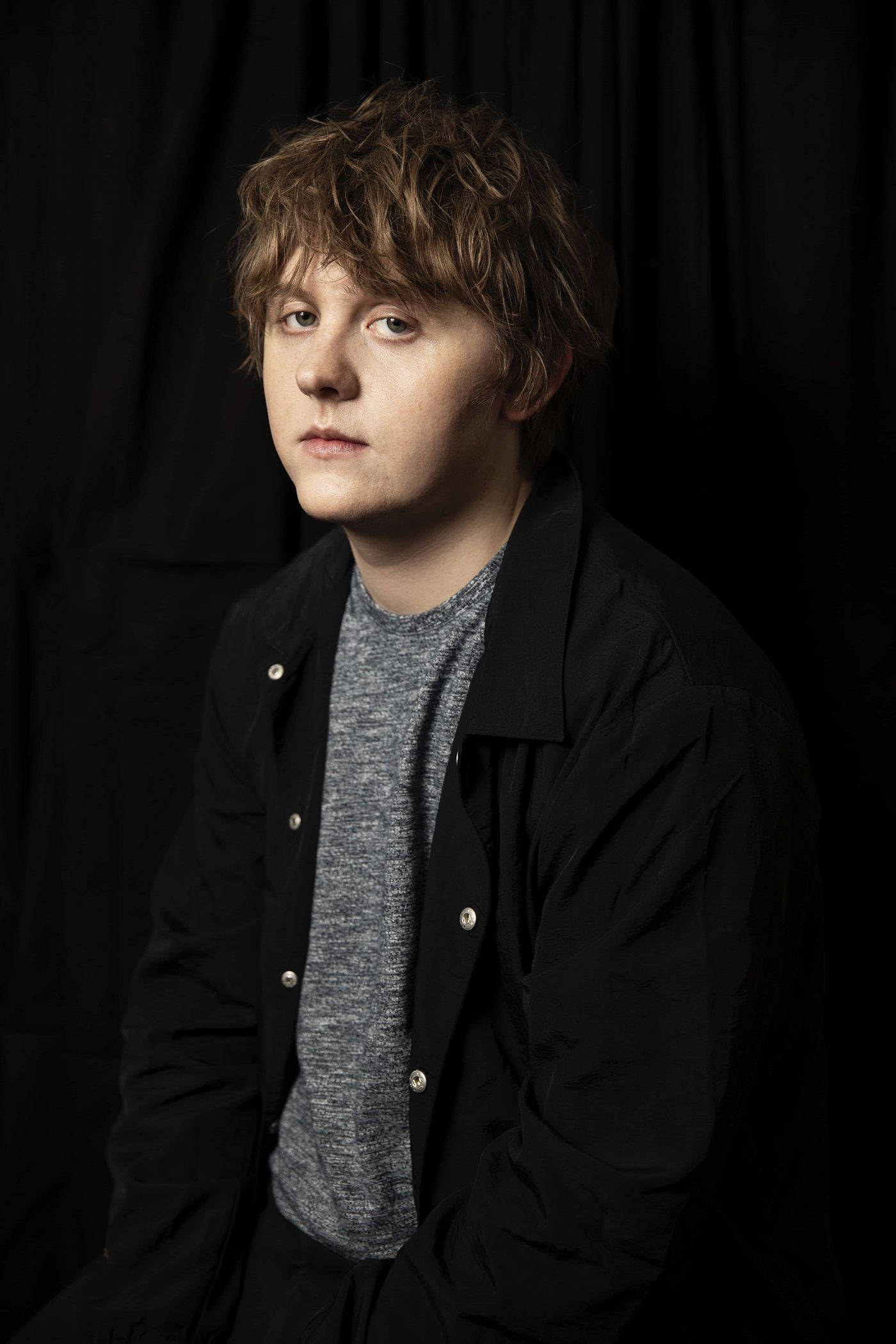 Lewis Capaldi 黑色背景照 壁紙 — 一位頭髮蓬鬆的年輕男子在鮮明的黑色背景下，若有所思地凝視著，營造出充滿氛圍的攝影棚肖像。