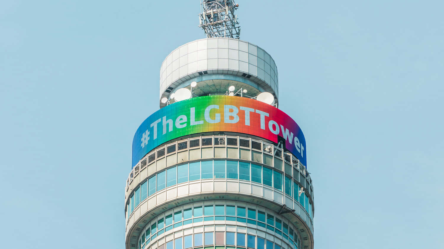 LGBT BT塔 壁紙 — 一座現代塔樓在清澈的藍天下展示帶有標籤 TheLGBTTower 的鮮豔彩虹橫幅。